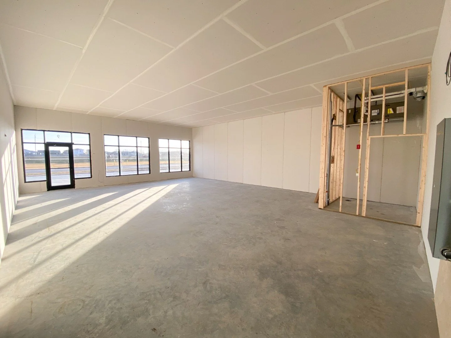 Interior of a new construction site for commercial space with large windows, concrete floor, and exposed wall framing near the right corner.