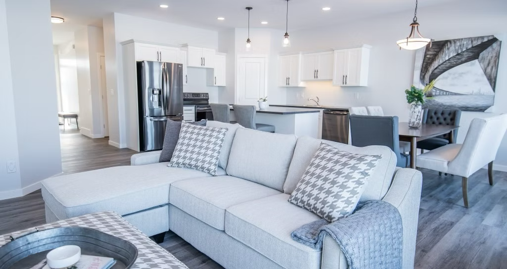 New development residential property open-concept living room and kitchen with white cabinets, stainless steel appliances, gray chairs, a dining table, and contemporary art.