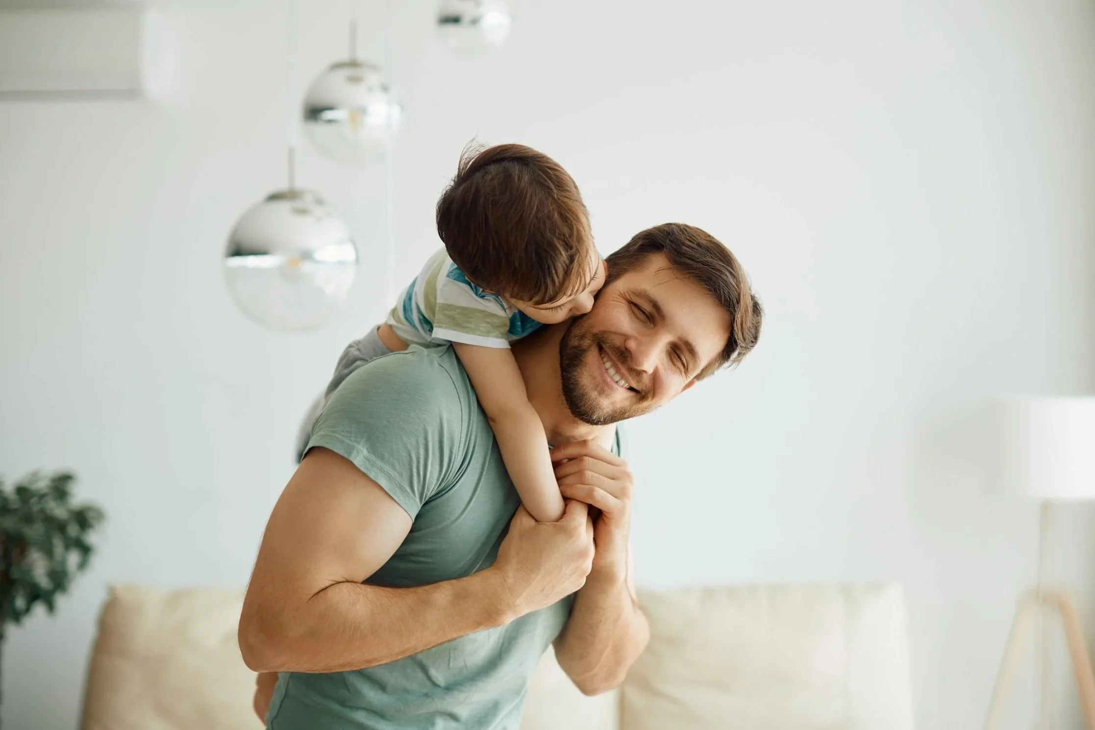 A man giving a piggyback ride to a young boy, both smiling happily indoors.