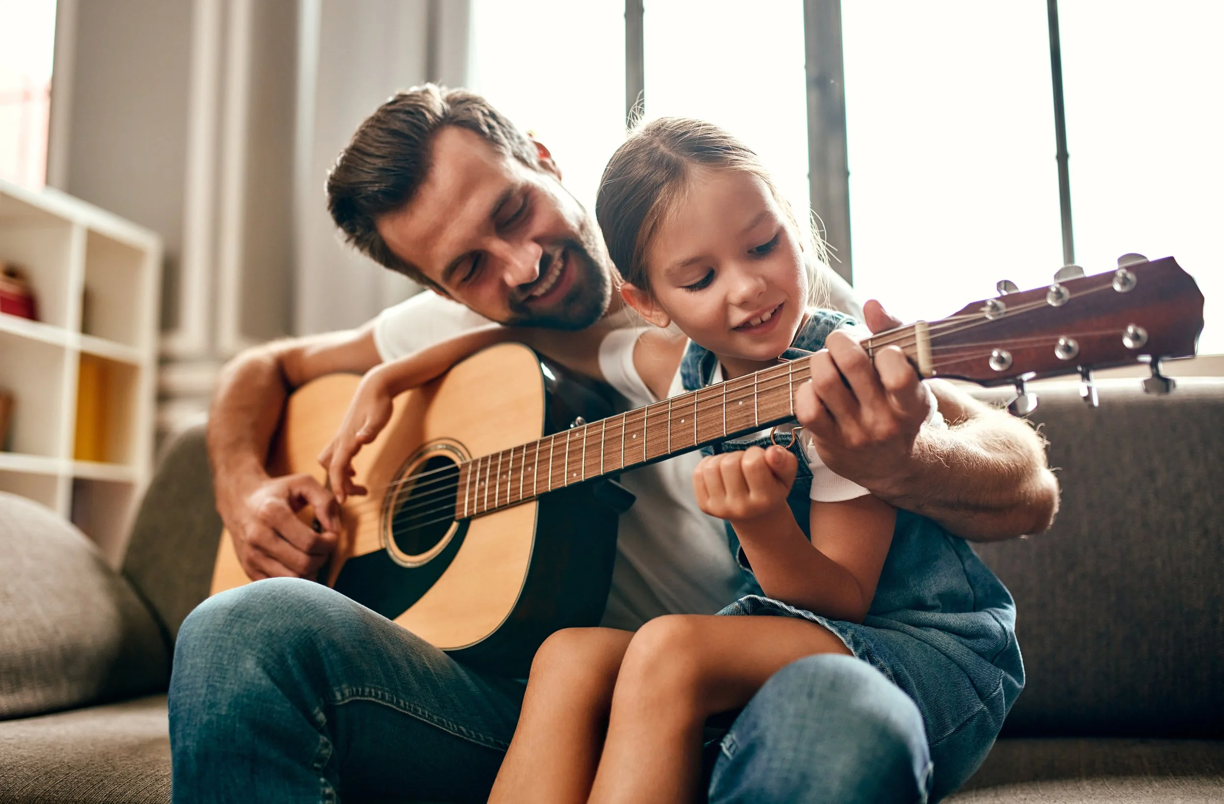 A man and a young girl sitting on a sofa playing guitar together in a well-lit living room.