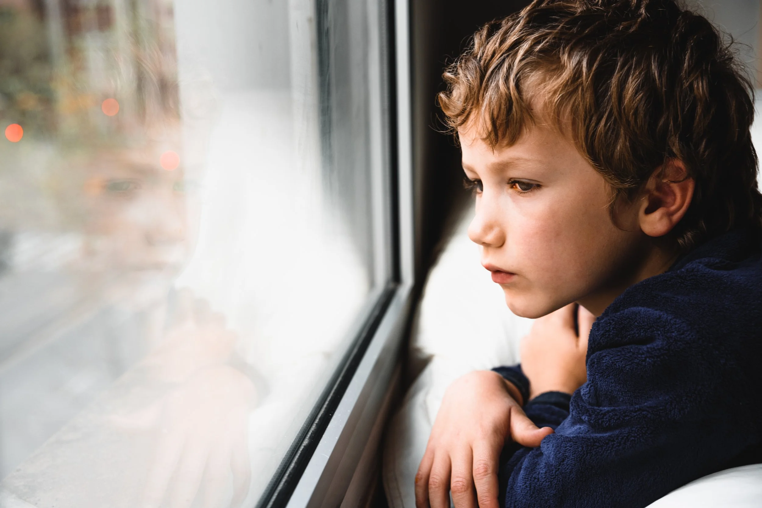 A young boy with curly brown hair looking out the window with a thoughtful expression, resting his arms on the windowsill, during daytime.