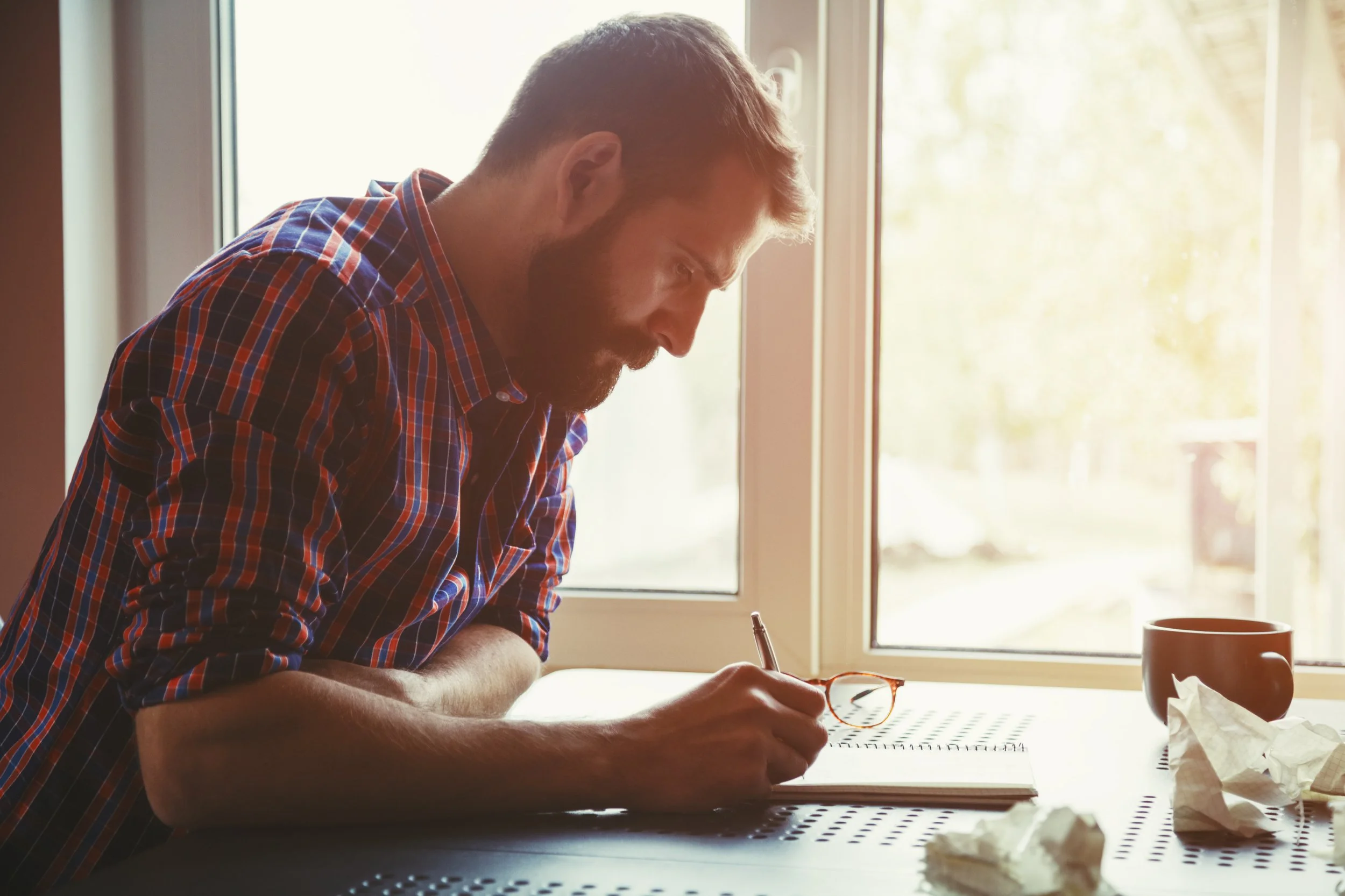 A man with glasses on a table, writing in a notebook near a window, with a mug and crumpled paper on the table.