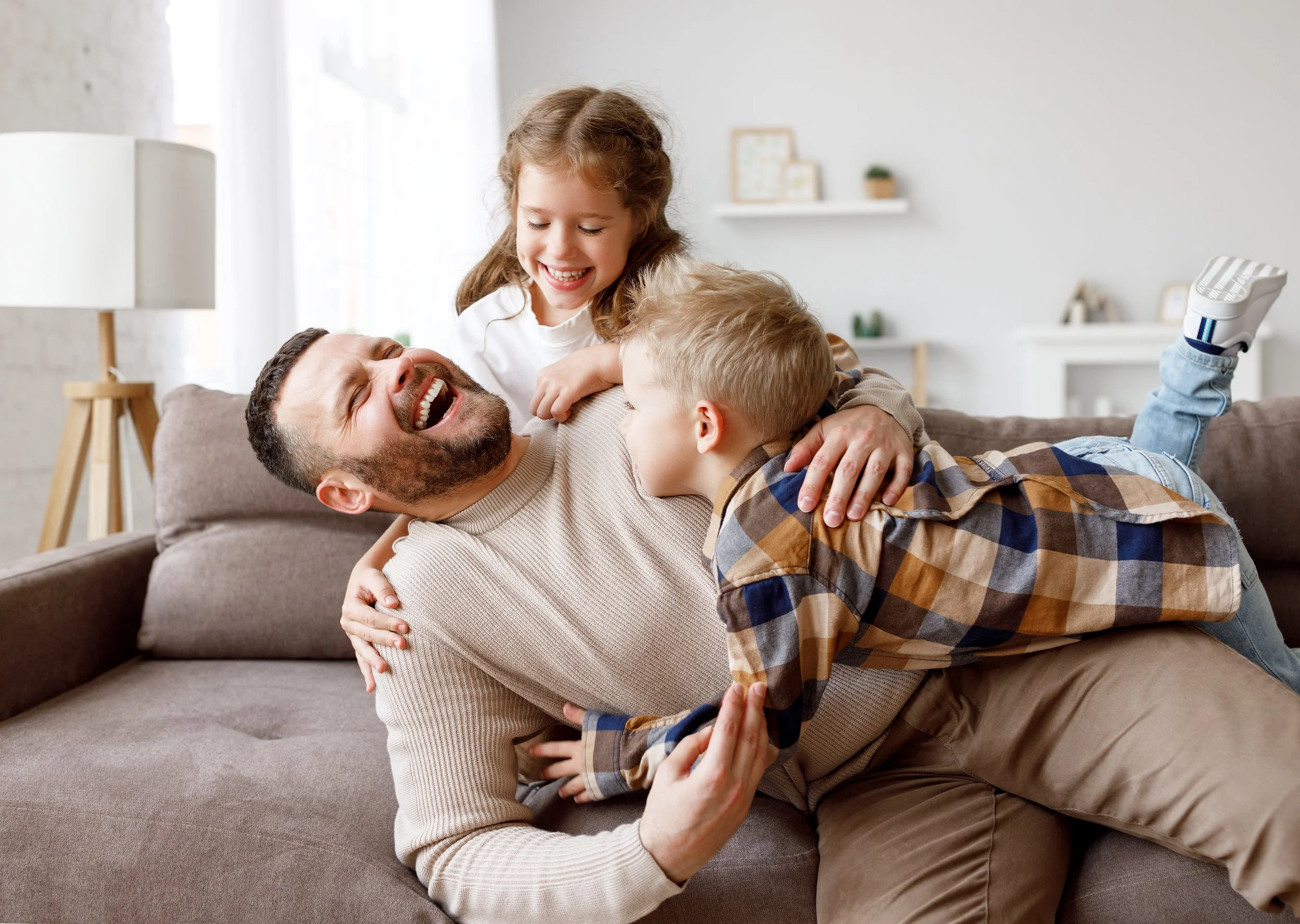Family of a father and two kids playing and laughing together on a couch in a living room.