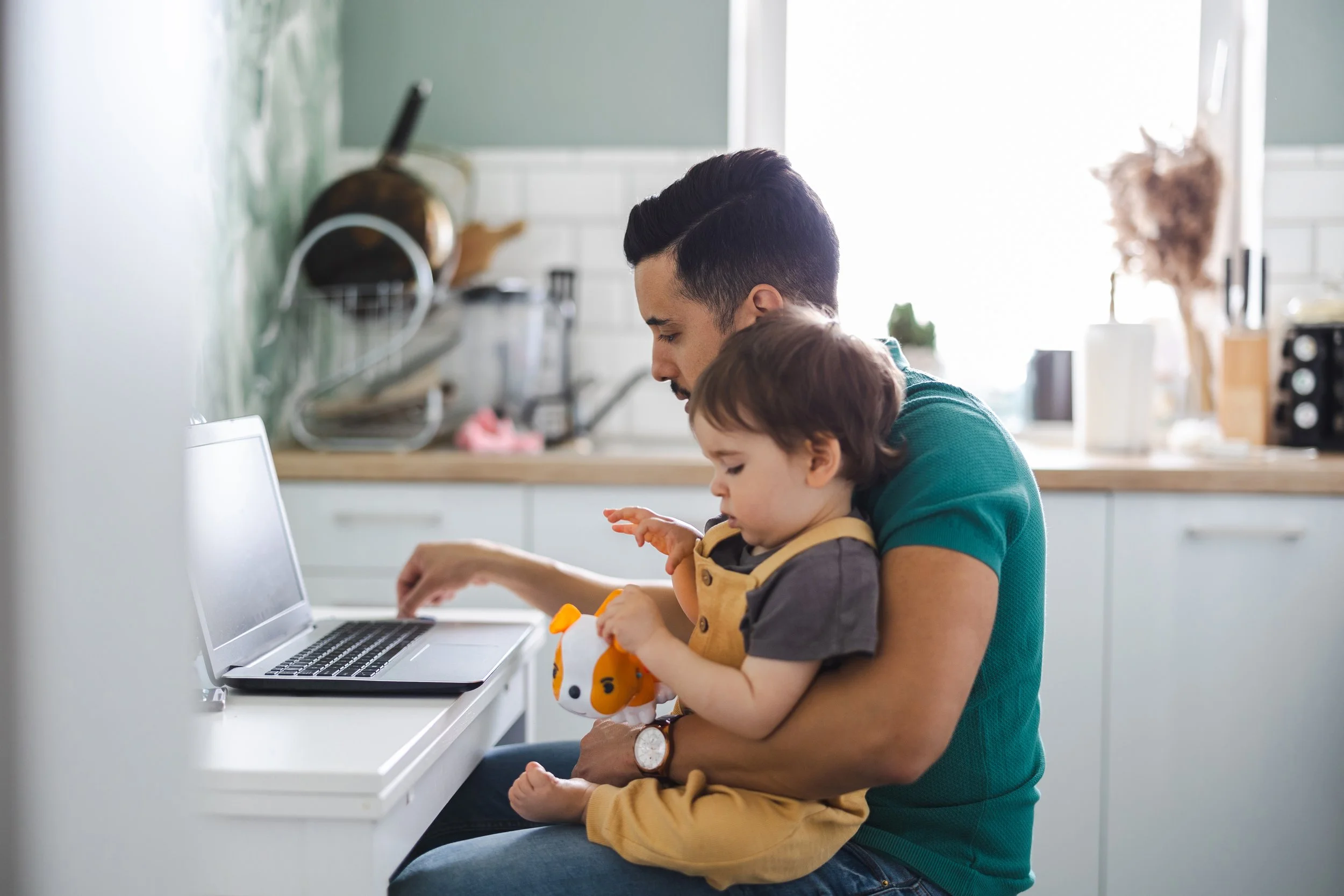 Man sitting on a chair with a young child on his lap while using a laptop in a kitchen.