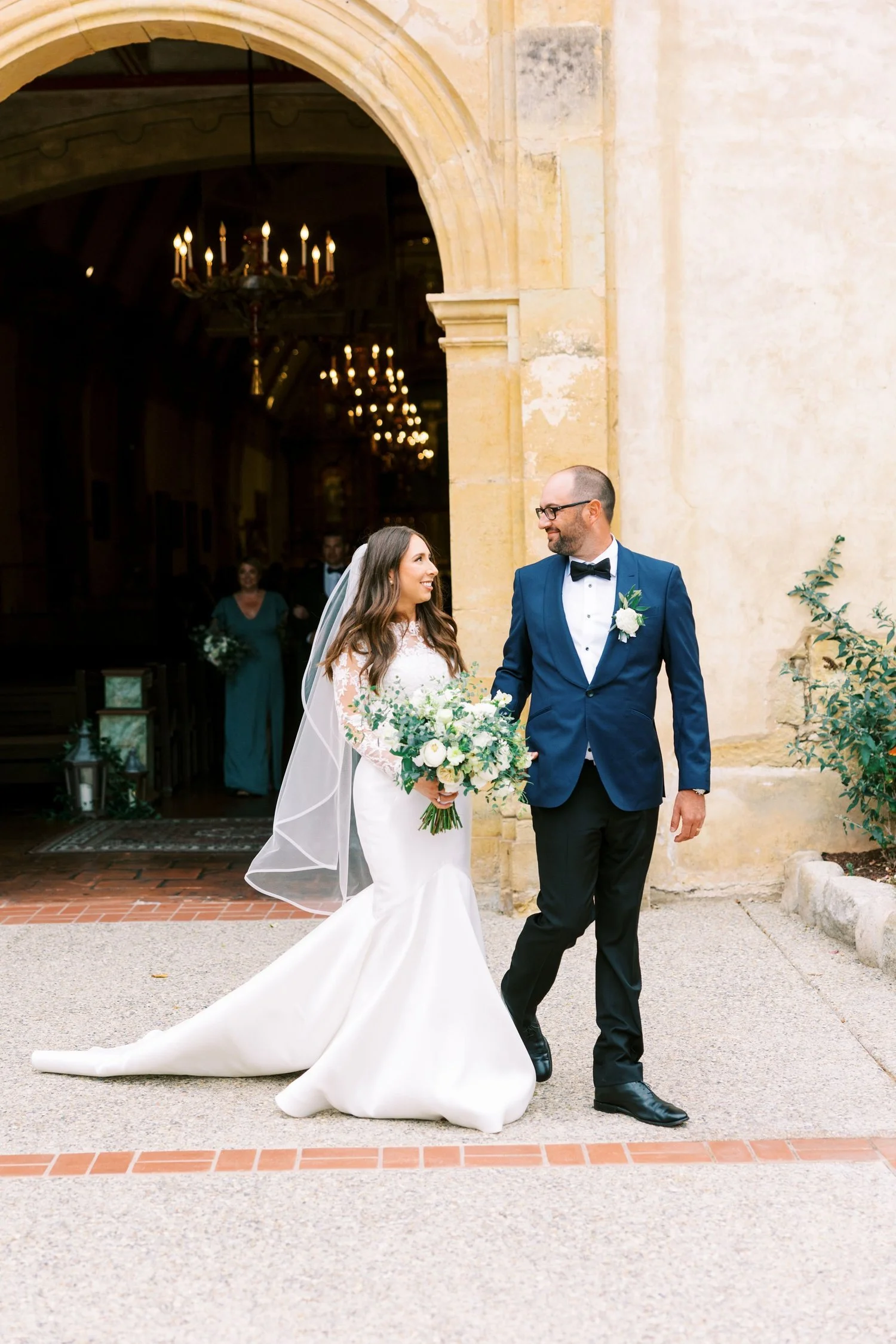Bride and groom walking out of a church, smiling at each other, with the bride holding a bouquet of white and green flowers.