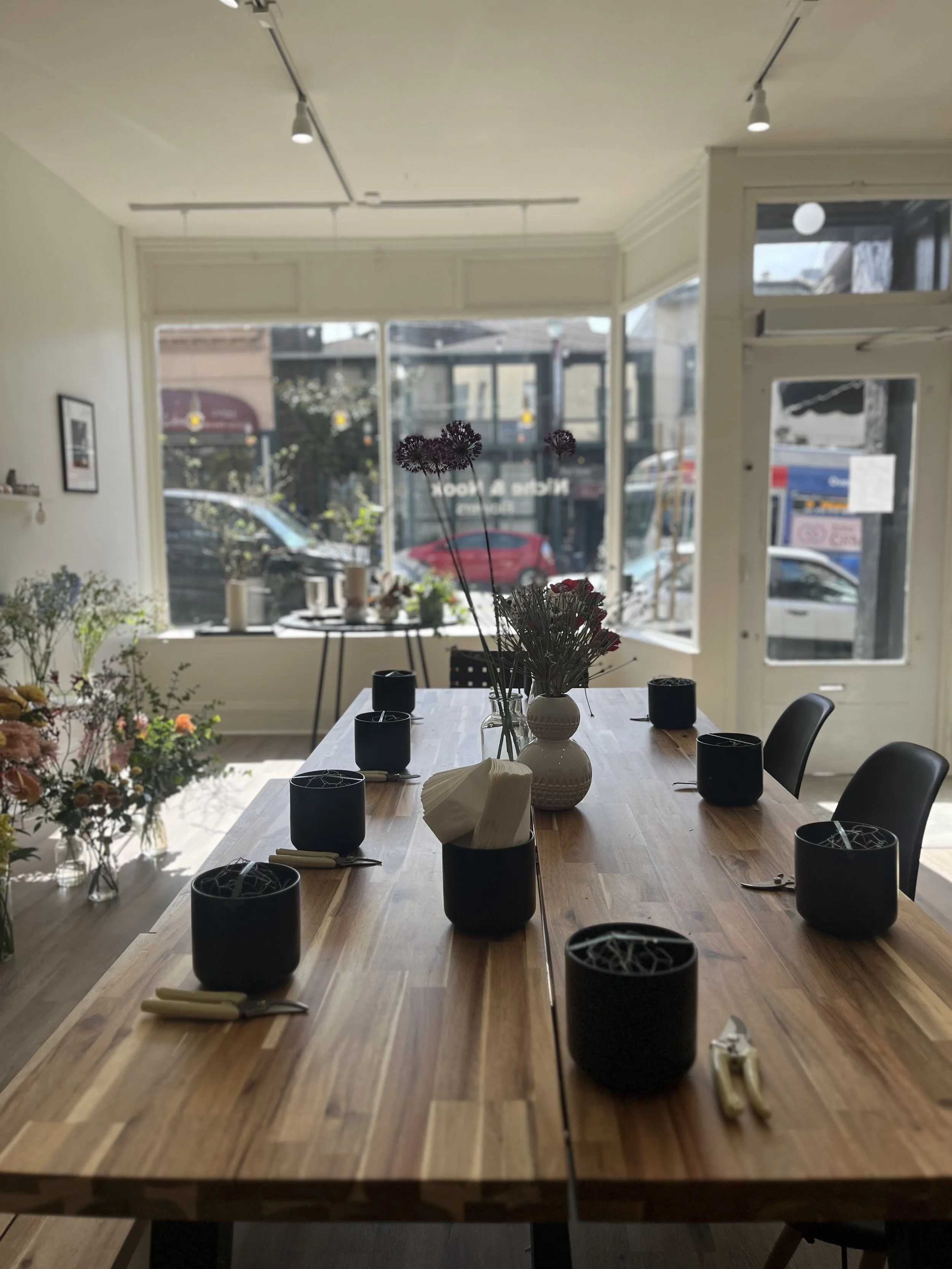 A wooden dining table set with black bowls, white napkins, and utensils, decorated with vases of flowers, inside a room with large front windows showing cars and buildings outside.