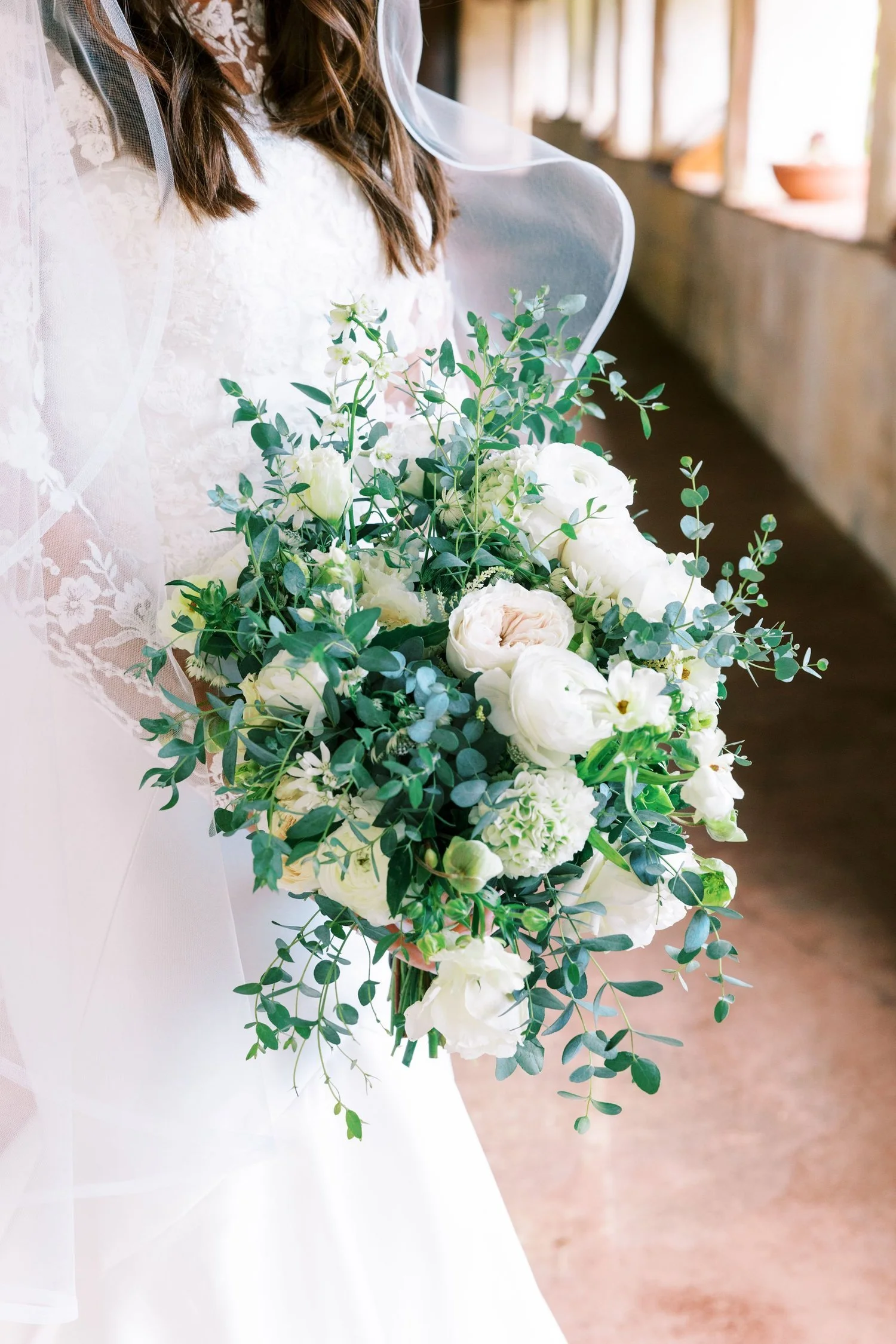 A bride in a lace wedding dress holding a large bouquet of white roses, peonies, and greenery.