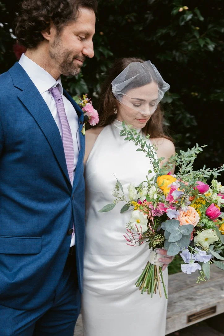 A bride and groom standing close together outdoors, with the groom in a blue suit and lavender tie, and the bride in a white gown with a veil, holding a colorful bouquet of flowers.