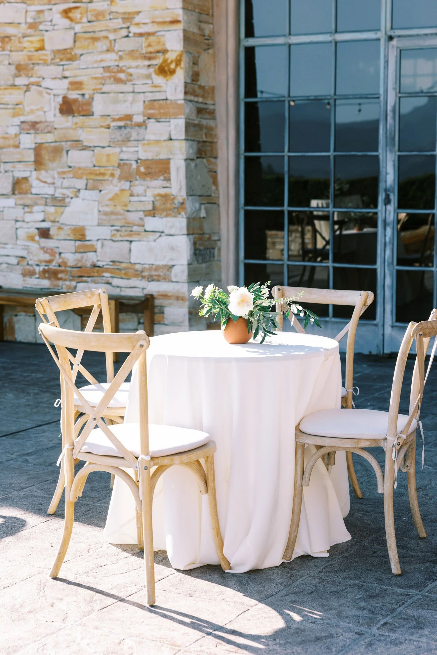 Outdoor table with a white tablecloth and a flower arrangement in a clay pot, surrounded by four wooden chairs with cushions, set against a brick wall and large window.