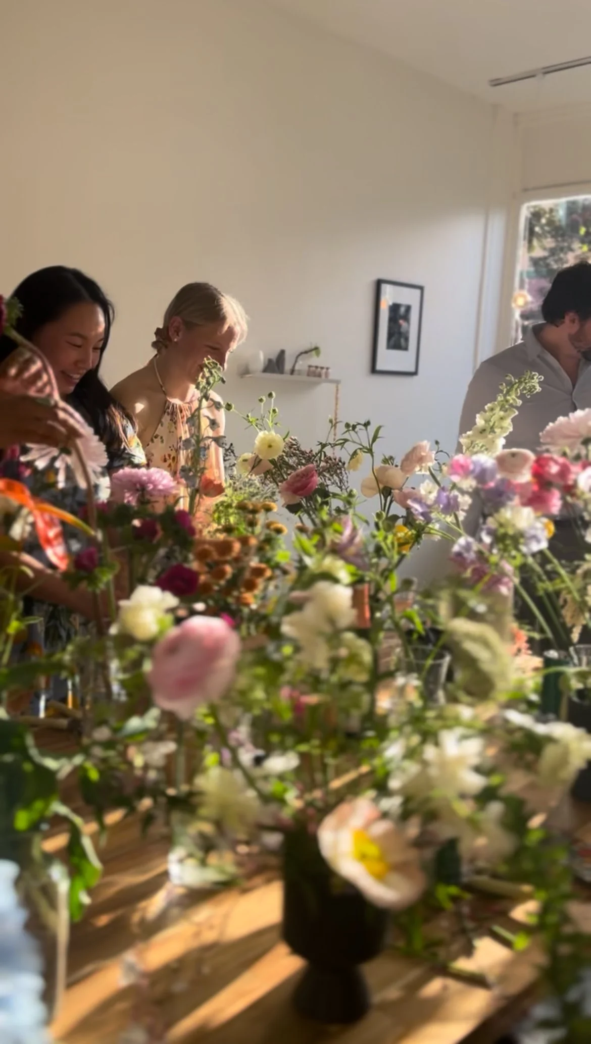 People smiling and looking at colorful flowers on a table, with a bright window in the background.