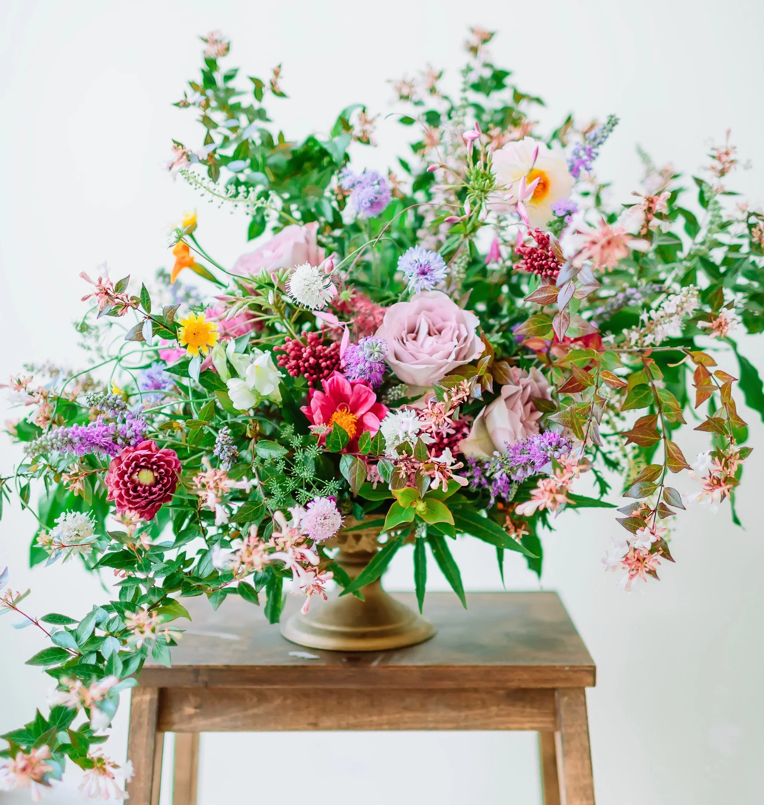 A large, colorful bouquet of mixed flowers in a vase on a wooden stool