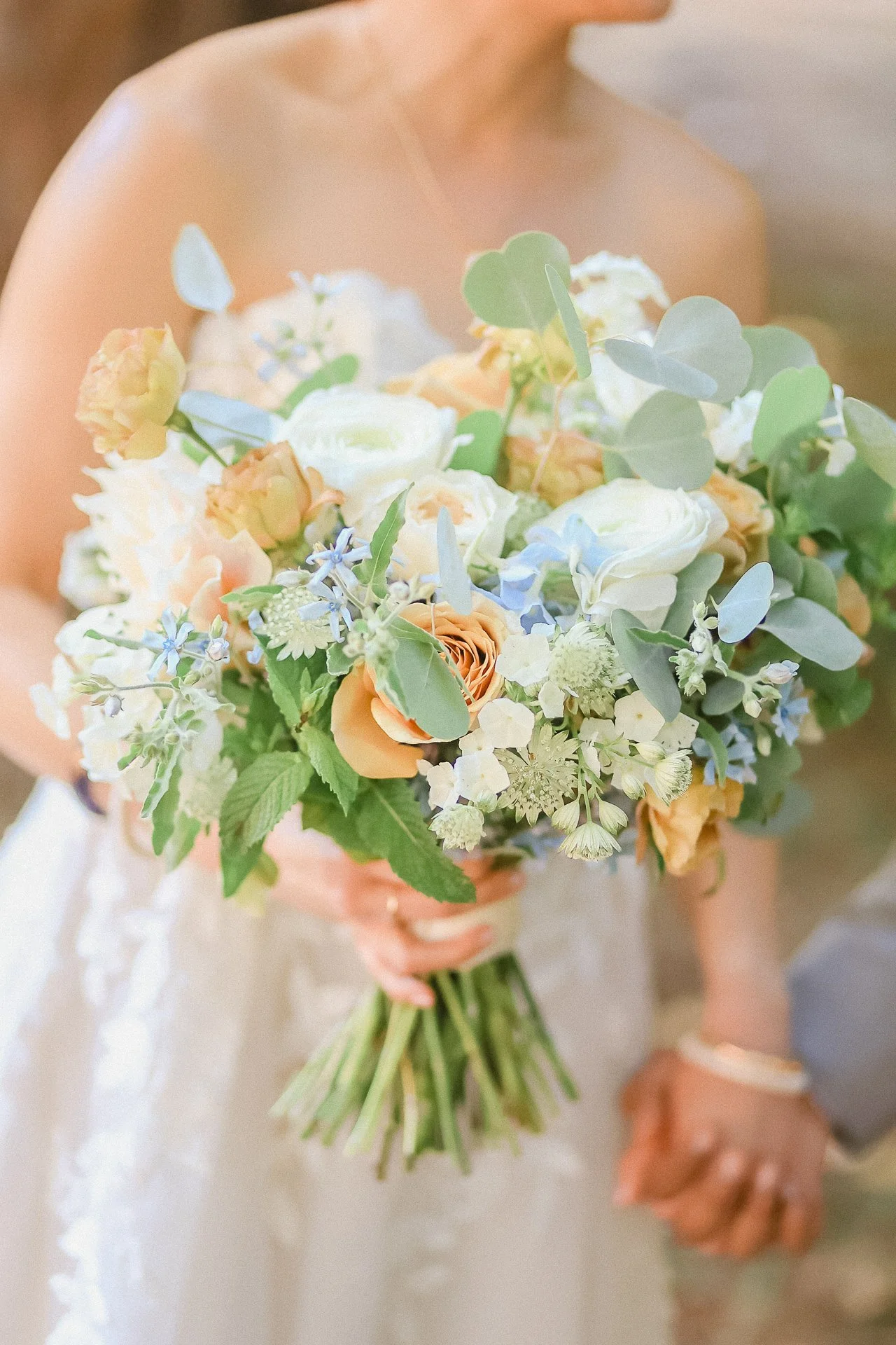 A bride holding a colorful bouquet of flowers with peach, white, off-white, blue, and green foliage, close-up.