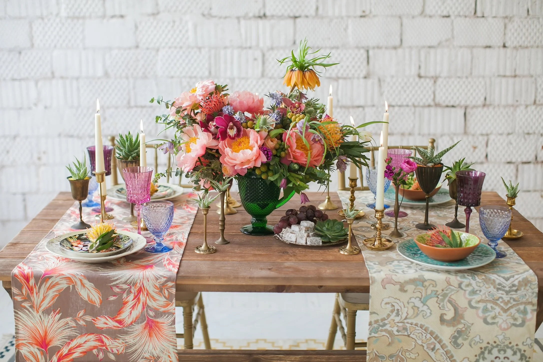 A wooden table decorated with colorful tableware, glassware, candles, and a large floral centerpiece, set against a white brick wall.