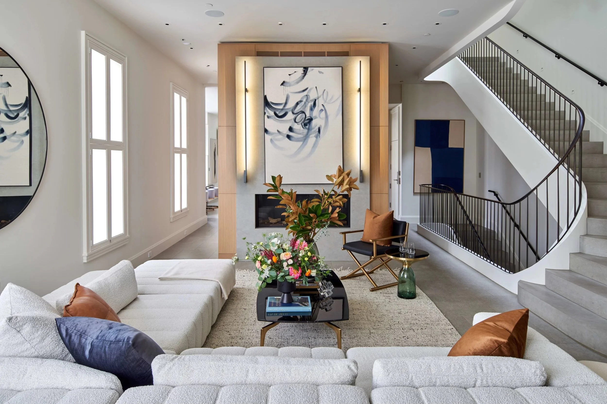 Modern living room with white sectional sofa, colorful throw pillows, black coffee table with books and a flower arrangement, armchair, glass side table with glasses, staircase with black railing, abstract art on walls, and a fireplace with large artwork above it.
