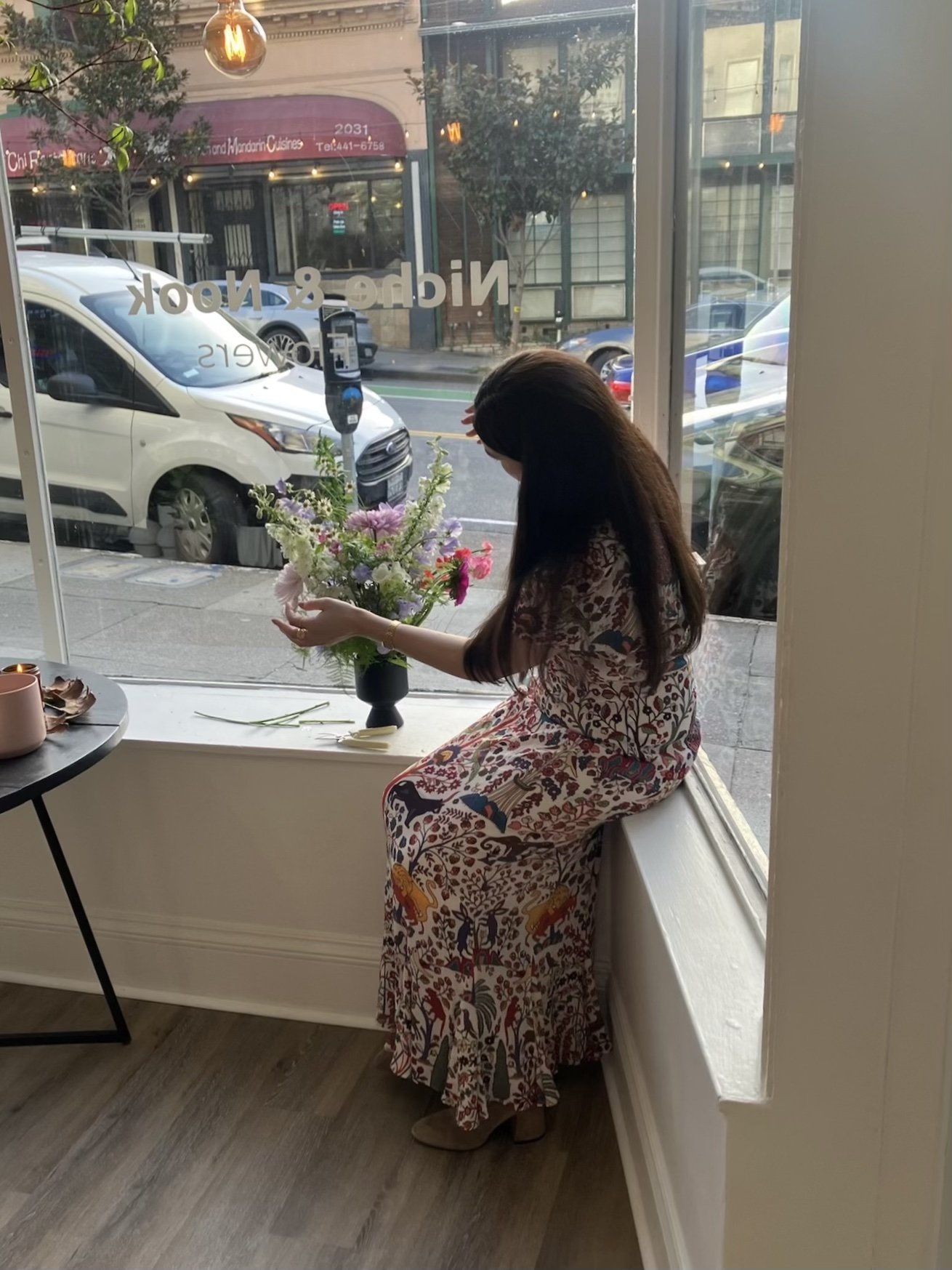 Woman sitting on window sill arranging a bouquet of flowers inside a café.