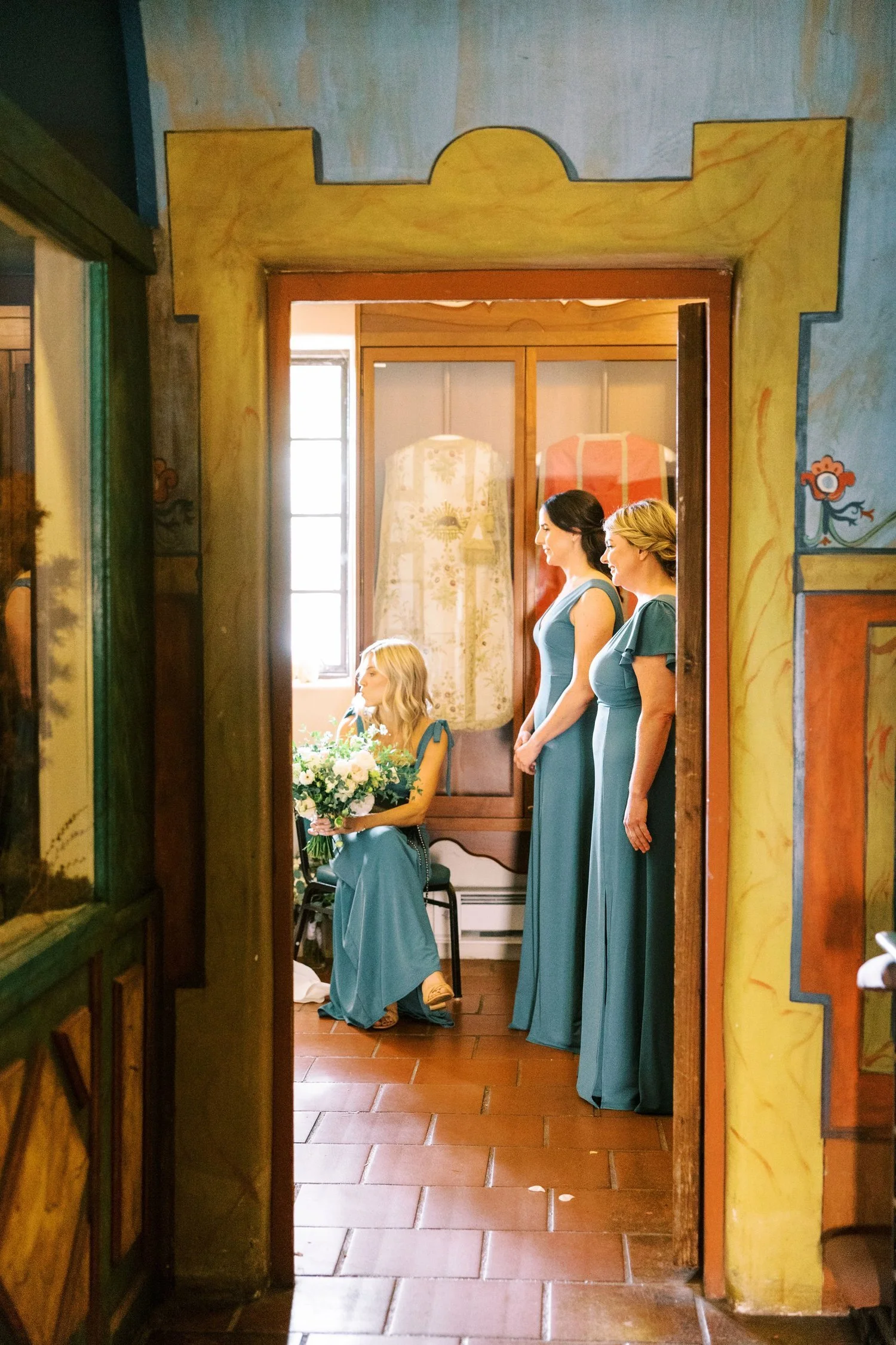 Four women in blue dresses, one sitting with a bouquet of flowers, three standing and facing her, inside a room with colorful painted walls and a window.