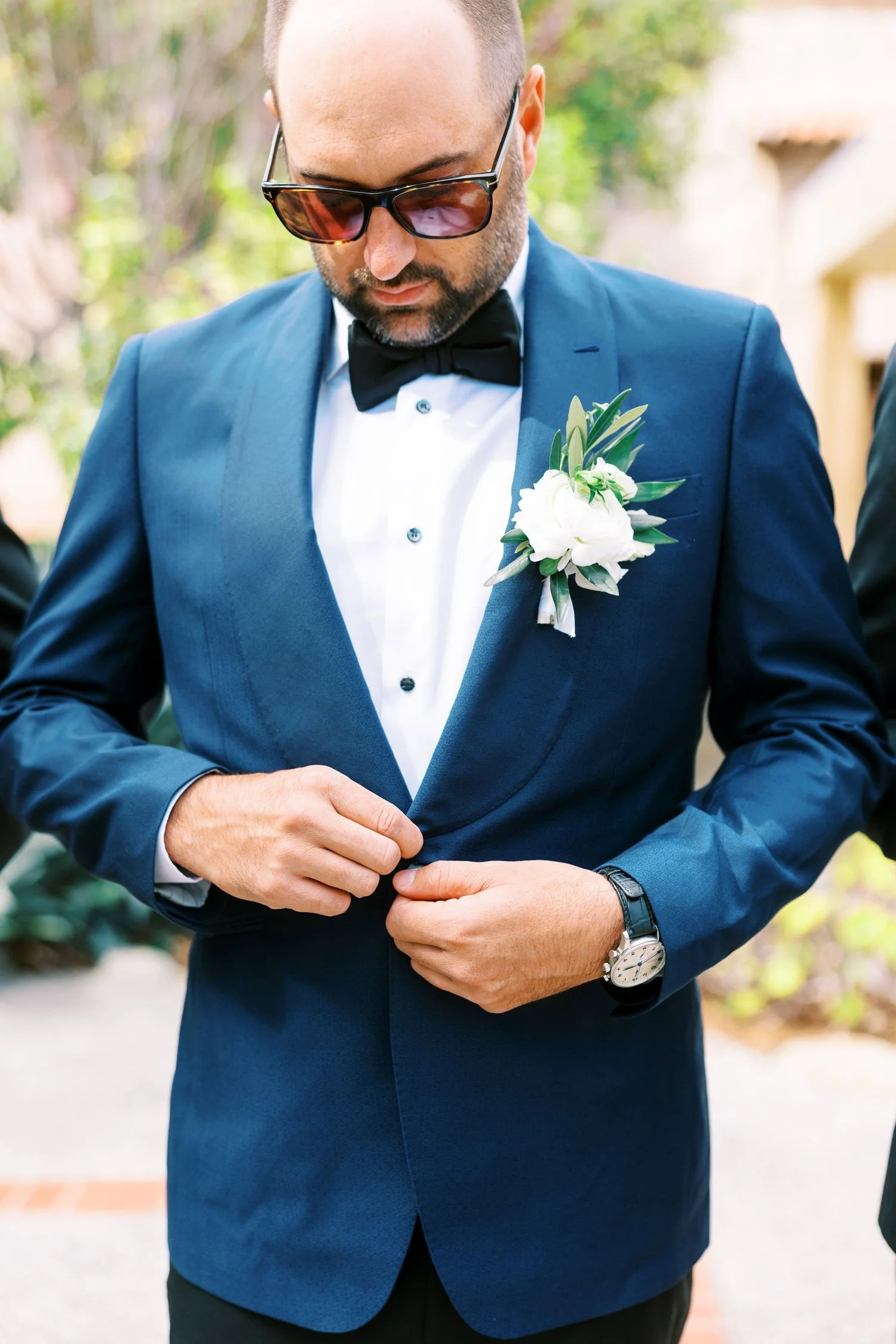 Man in a blue tuxedo with a boutonnière, adjusting his jacket at an outdoor event.