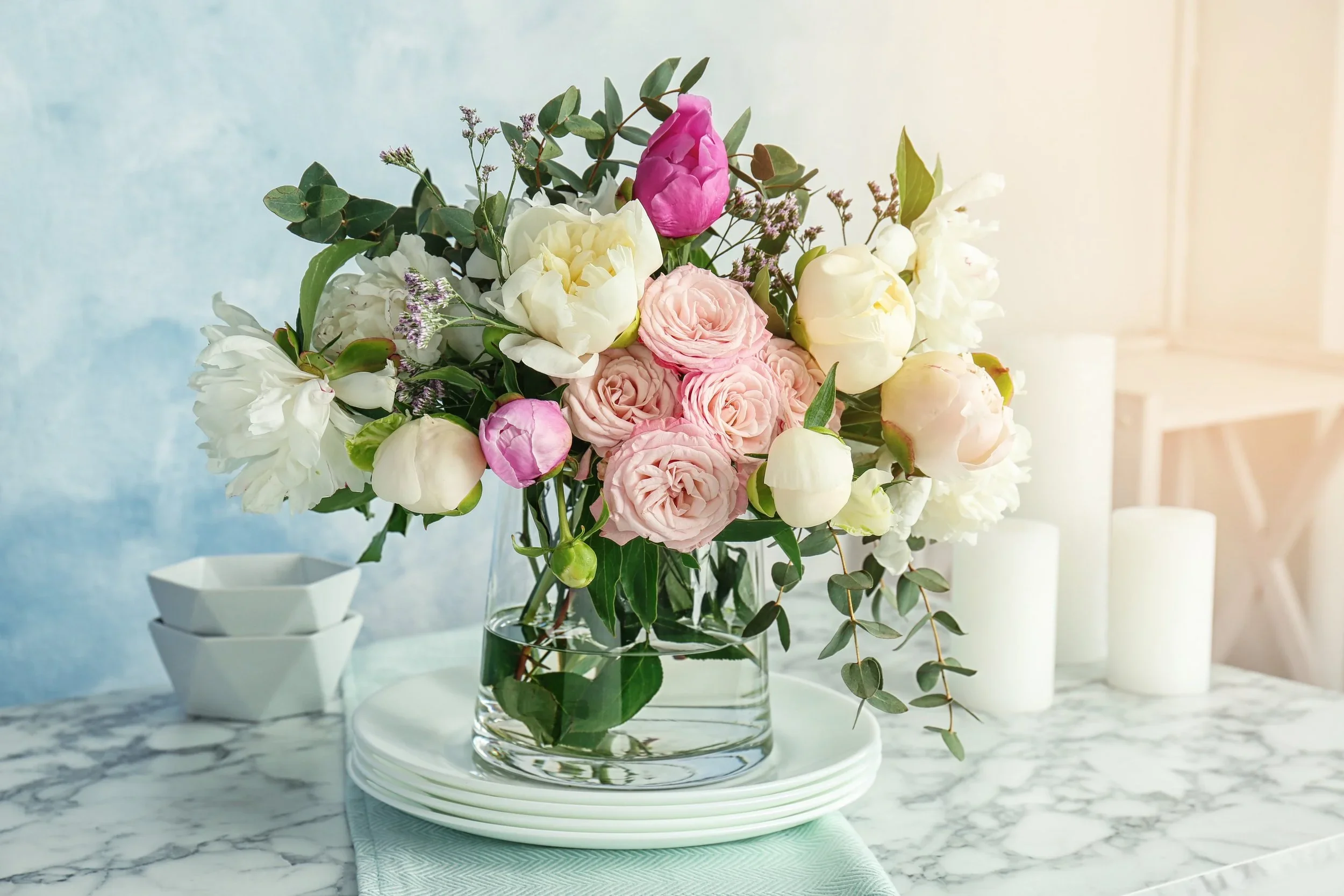 A bouquet of pink, white, and purple flowers in a glass vase on a marble table with white candles and bowls in the background.