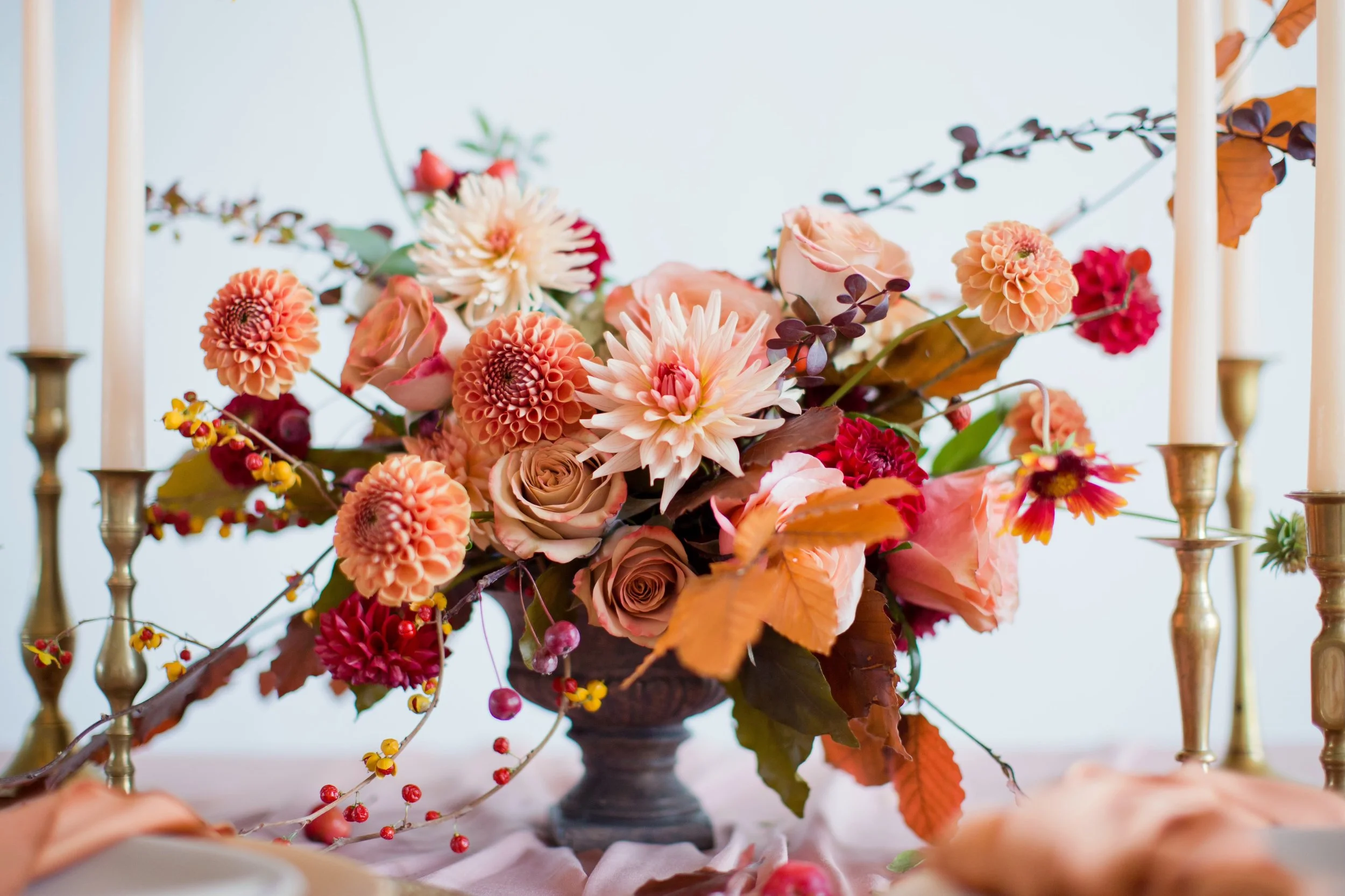 A floral arrangement with peaches, pink, and red dahlias, roses, and autumn leaves in a black vase, flanked by tall candlesticks.