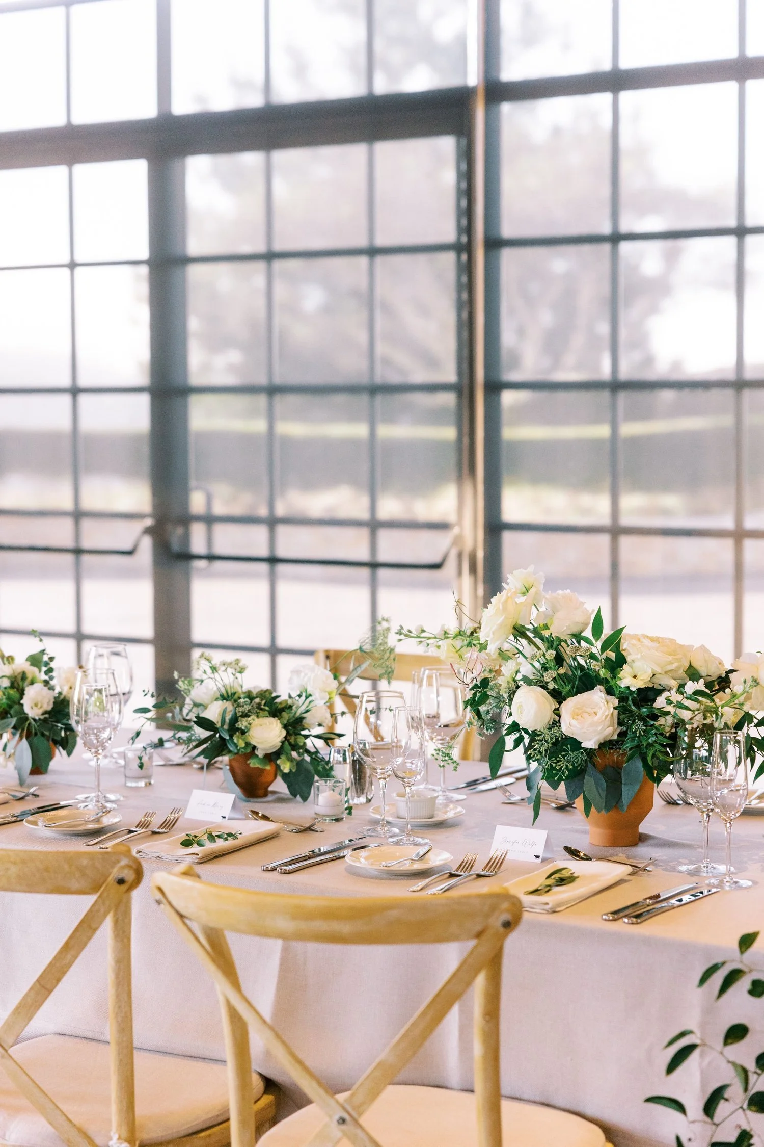 Elegant banquet table set for a formal event with white floral arrangements, glassware, and cutlery, in a bright room with large grid-style windows.