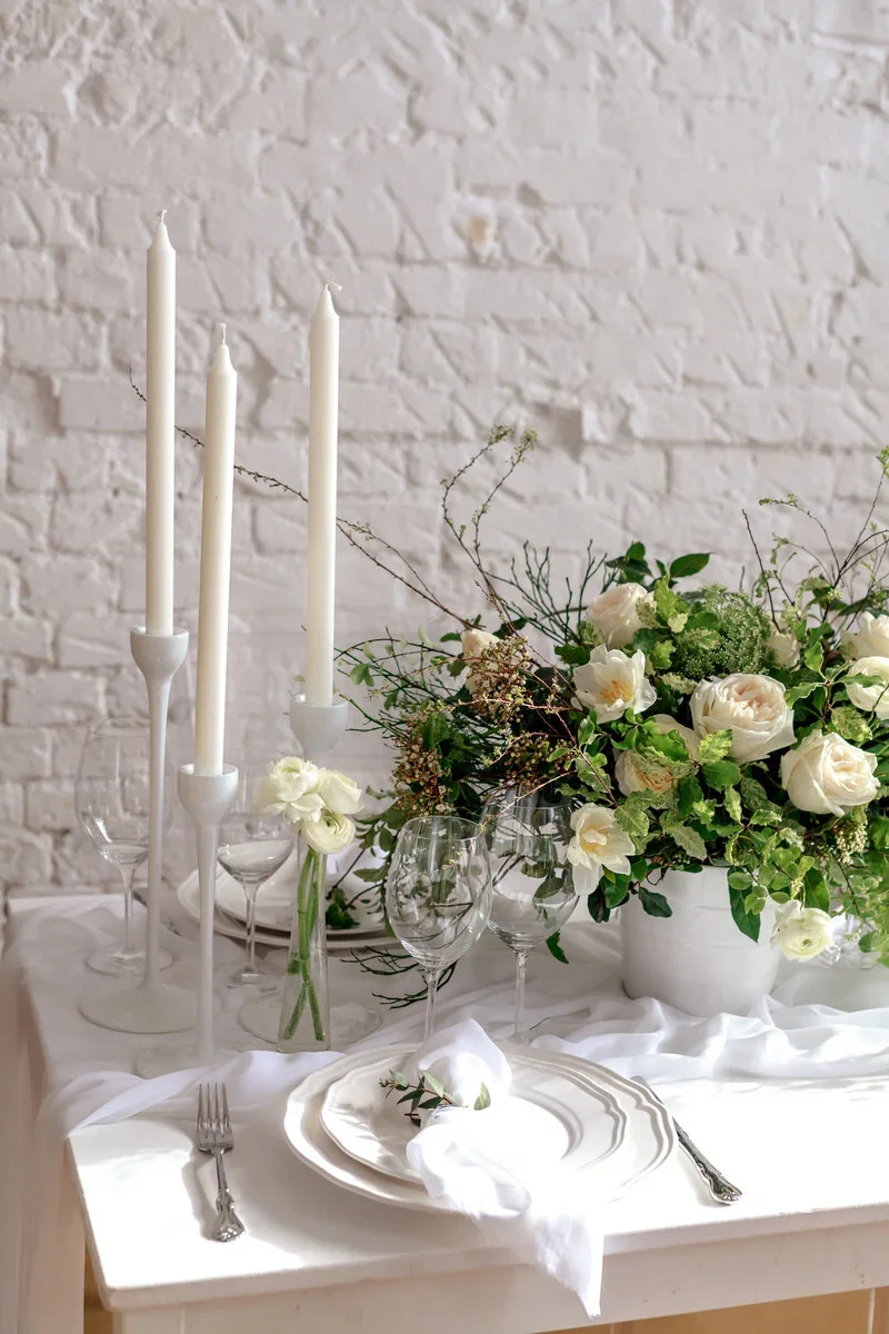 Elegant table setting with white plates, silverware, a white cloth napkin, tall white candles in candlesticks, a glass with white flowers, and a large arrangement of white roses and greenery in a white vase, against a white brick wall.