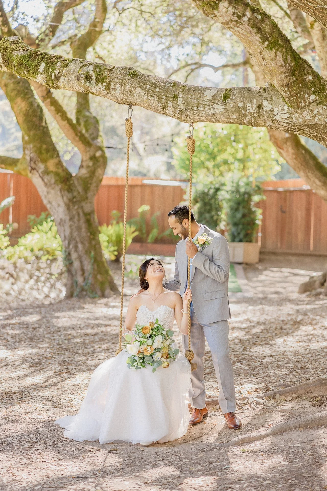 A couple on their wedding day with the bride sitting on a swing holding a bouquet of flowers, and the groom standing beside her, exchanging loving looks in a sunny outdoor setting with trees and a wooden fence in the background.