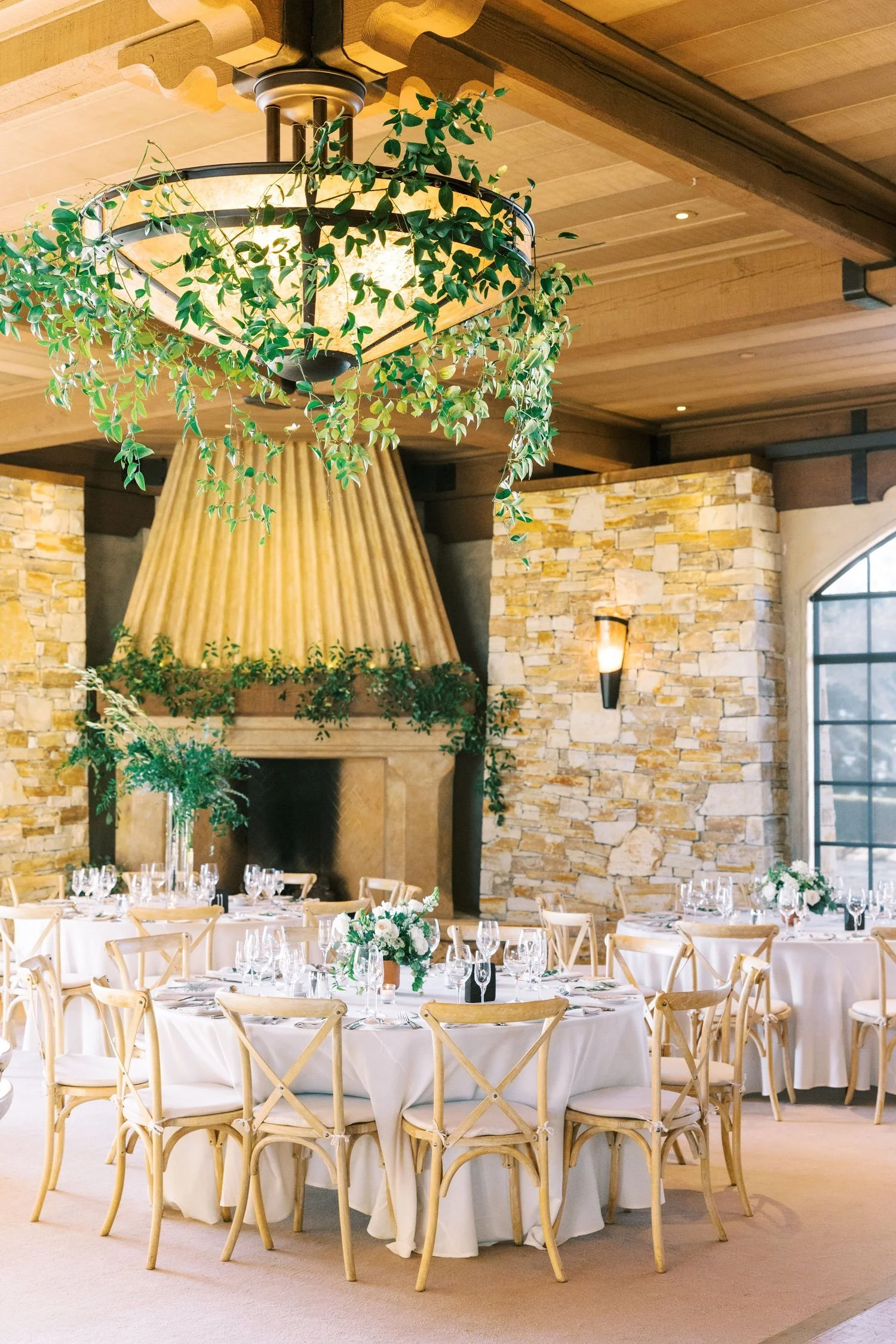 Elegant banquet hall decorated with round tables covered in white tablecloths, each set with wine glasses, plates, silverware, and floral centerpieces. The room features a large stone fireplace, warm wooden ceiling, hanging greenery, and a vintage ch