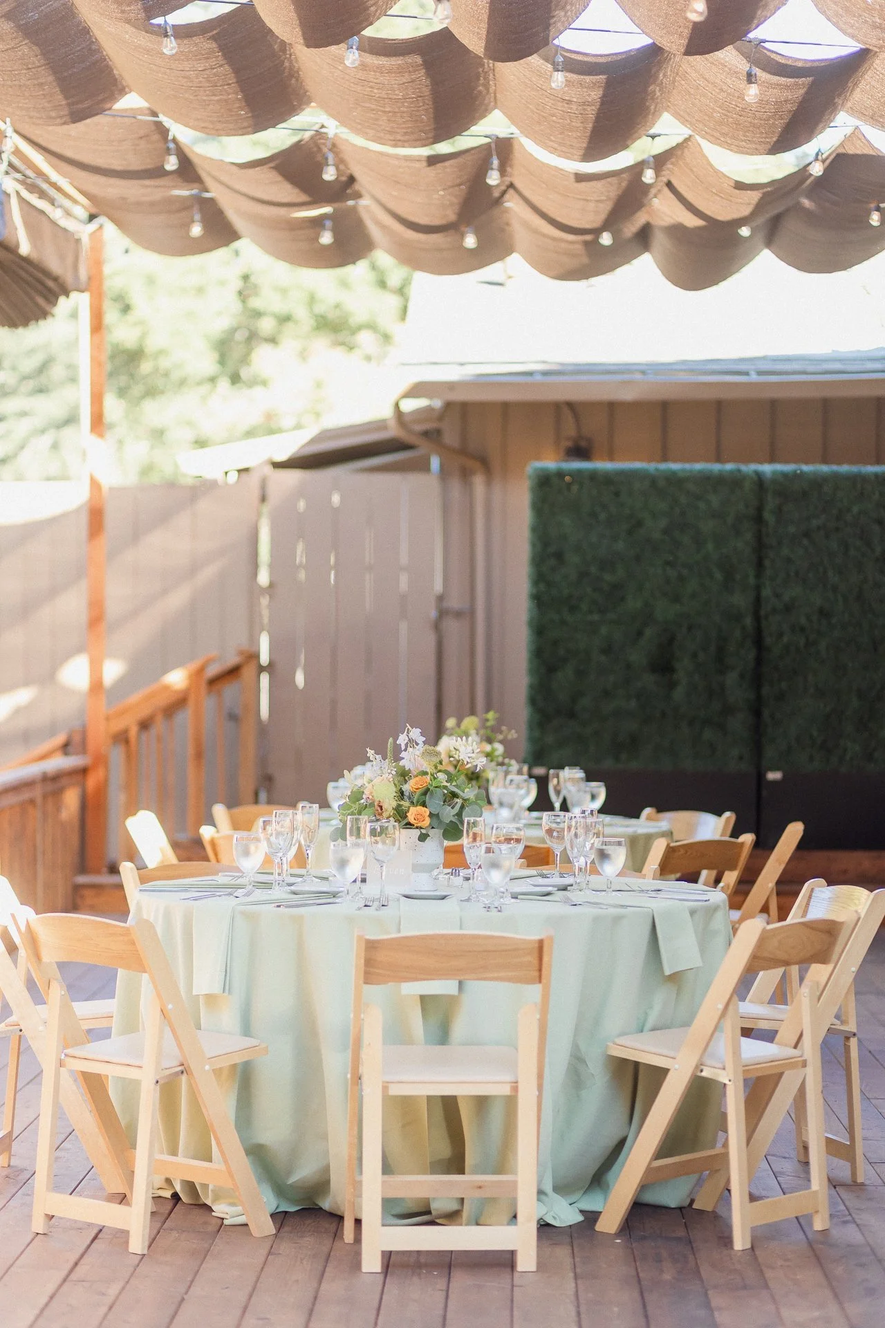Outdoor party setup with a round table covered in a light green tablecloth, decorated with a flower centerpiece, surrounded by wooden chairs, under a canopy of brown fabric and string lights.