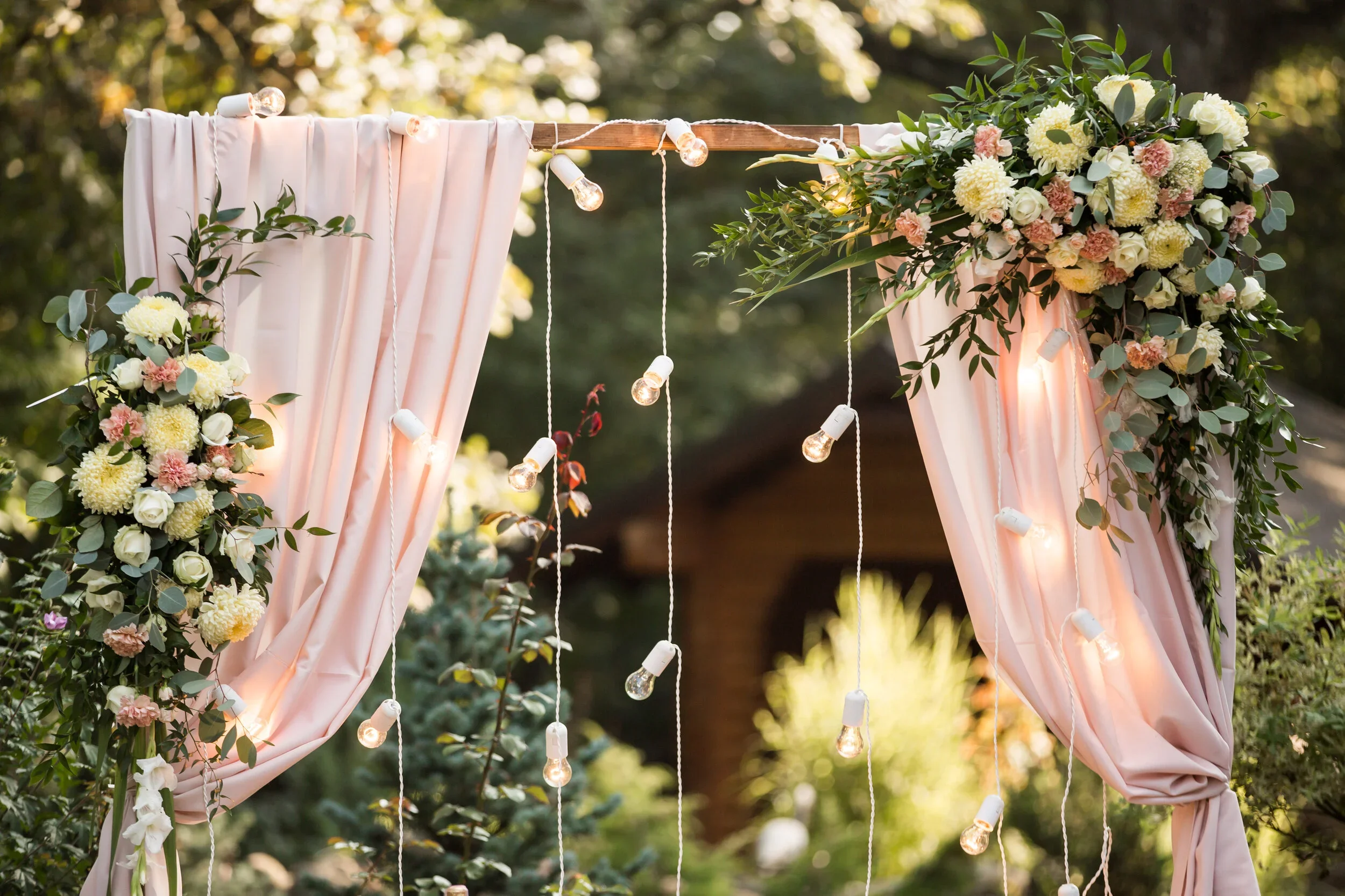 Decorative outdoor wedding arch with pink curtains, floral arrangements of white, cream, and pink flowers, greenery, and hanging string lights.