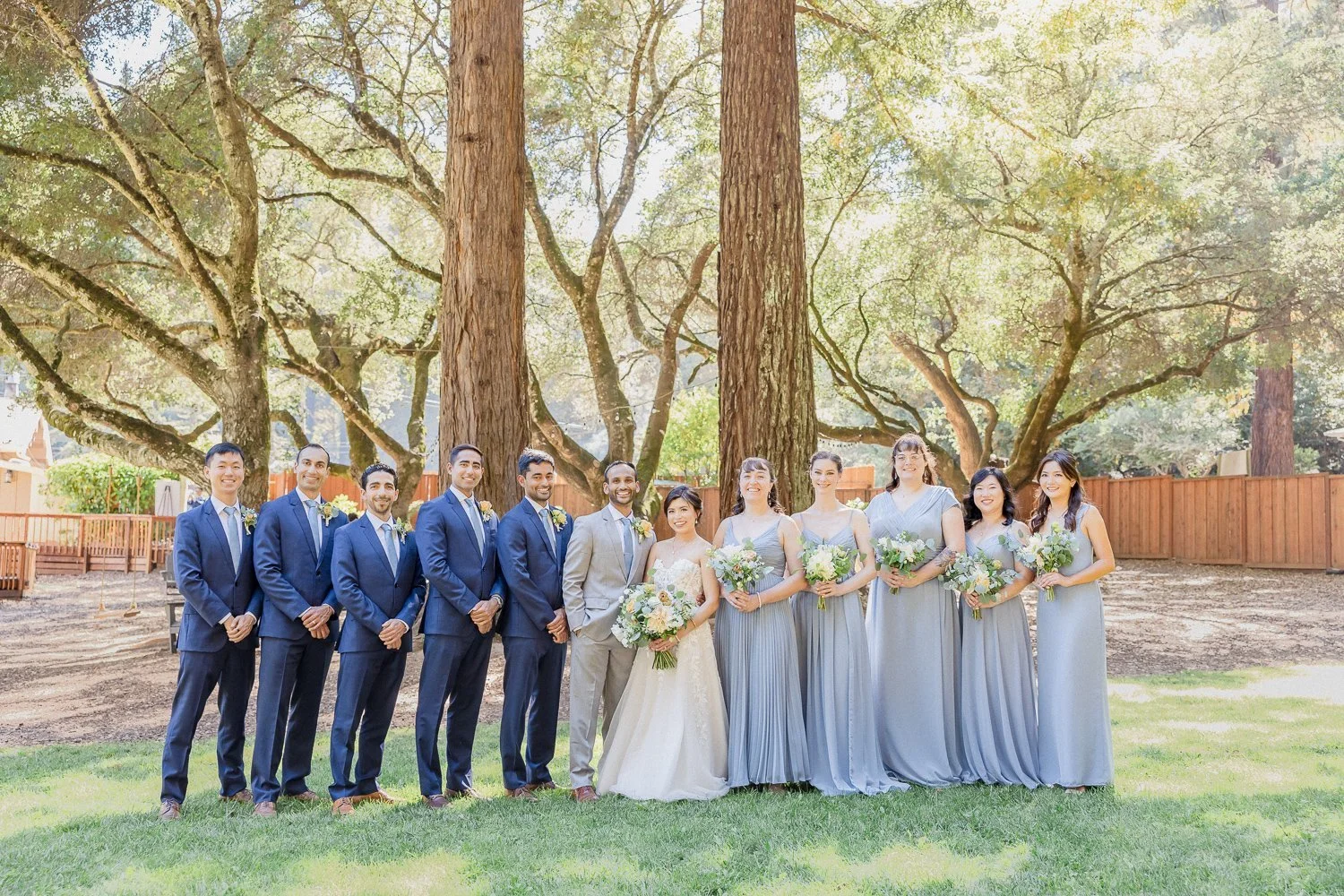 A wedding party standing outdoors in front of large trees, with the bride, groom, bridesmaids, and groomsmen all smiling and holding bouquets.