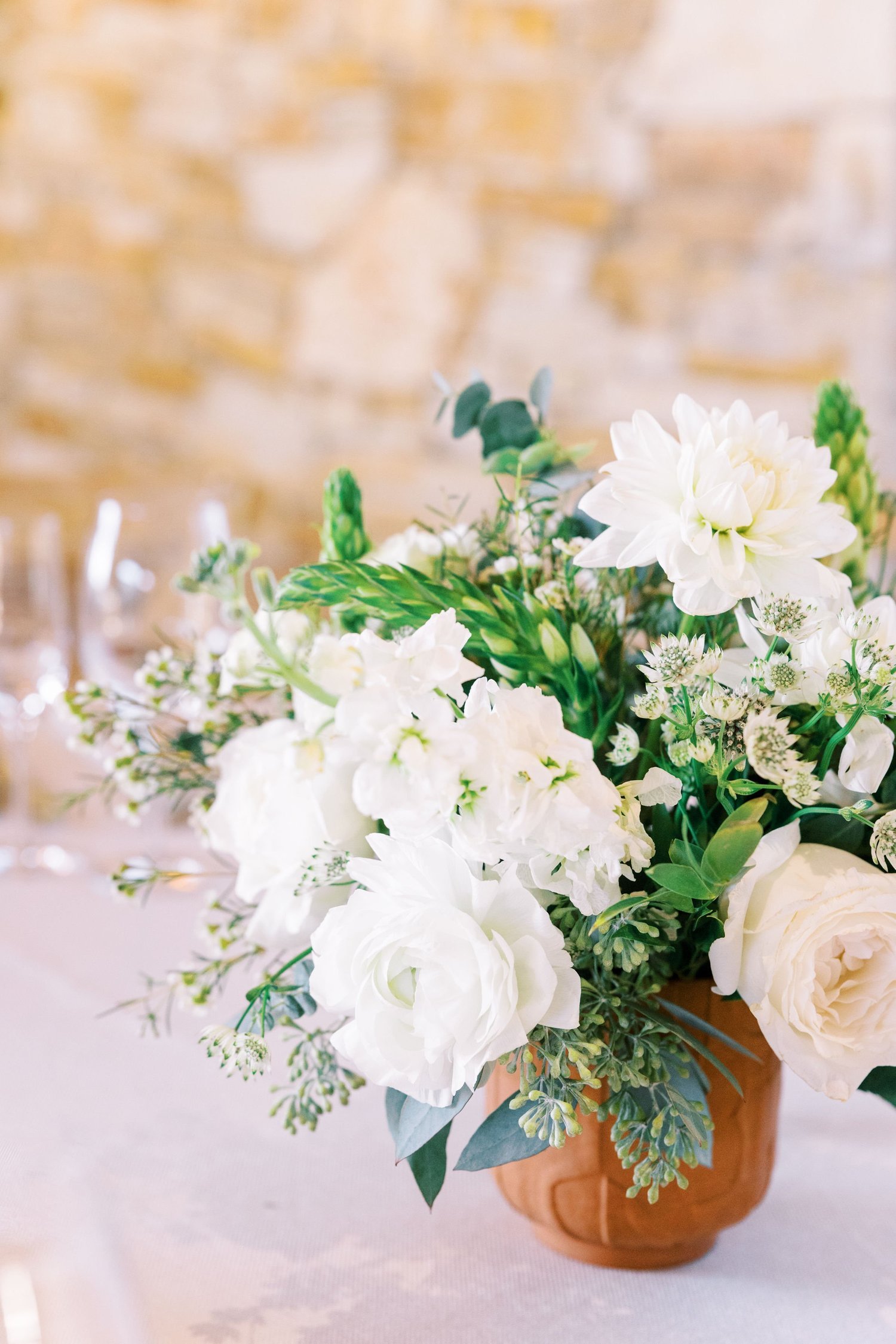 White floral arrangement in a terracotta pot on a table with a blurred brick wall background.