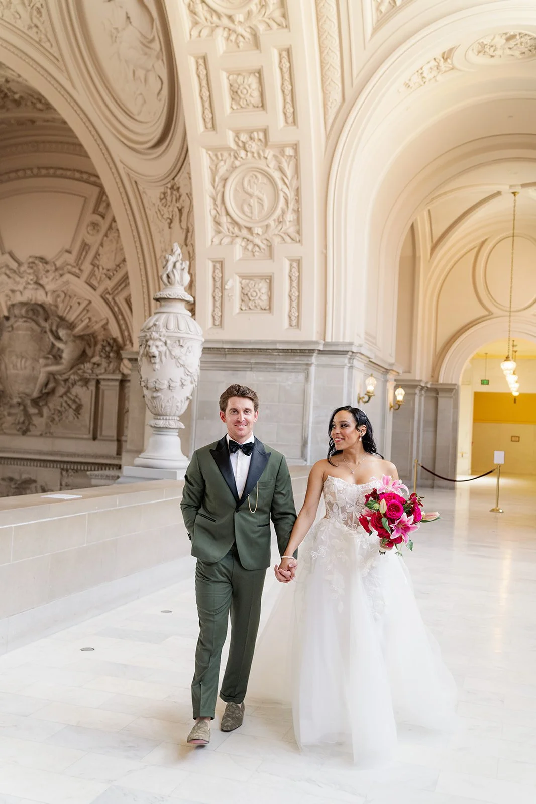 A bride and groom holding hands, walking in a grand, ornate hall with cream-colored walls and artistic sculptures.