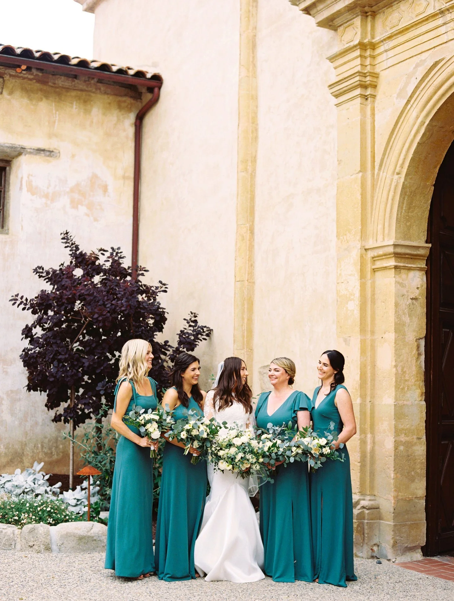 Bride and four bridesmaids standing outdoors in front of an old building, holding bouquets, all smiling and dressed in teal gowns.