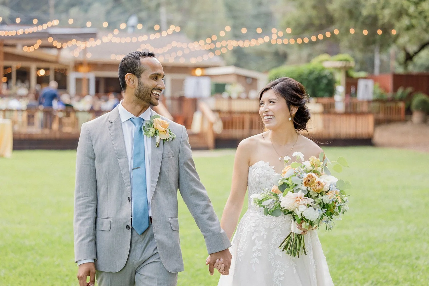 A newlywed couple holding hands and smiling at each other outdoors at sunset, with string lights and a gathering of guests in the background.