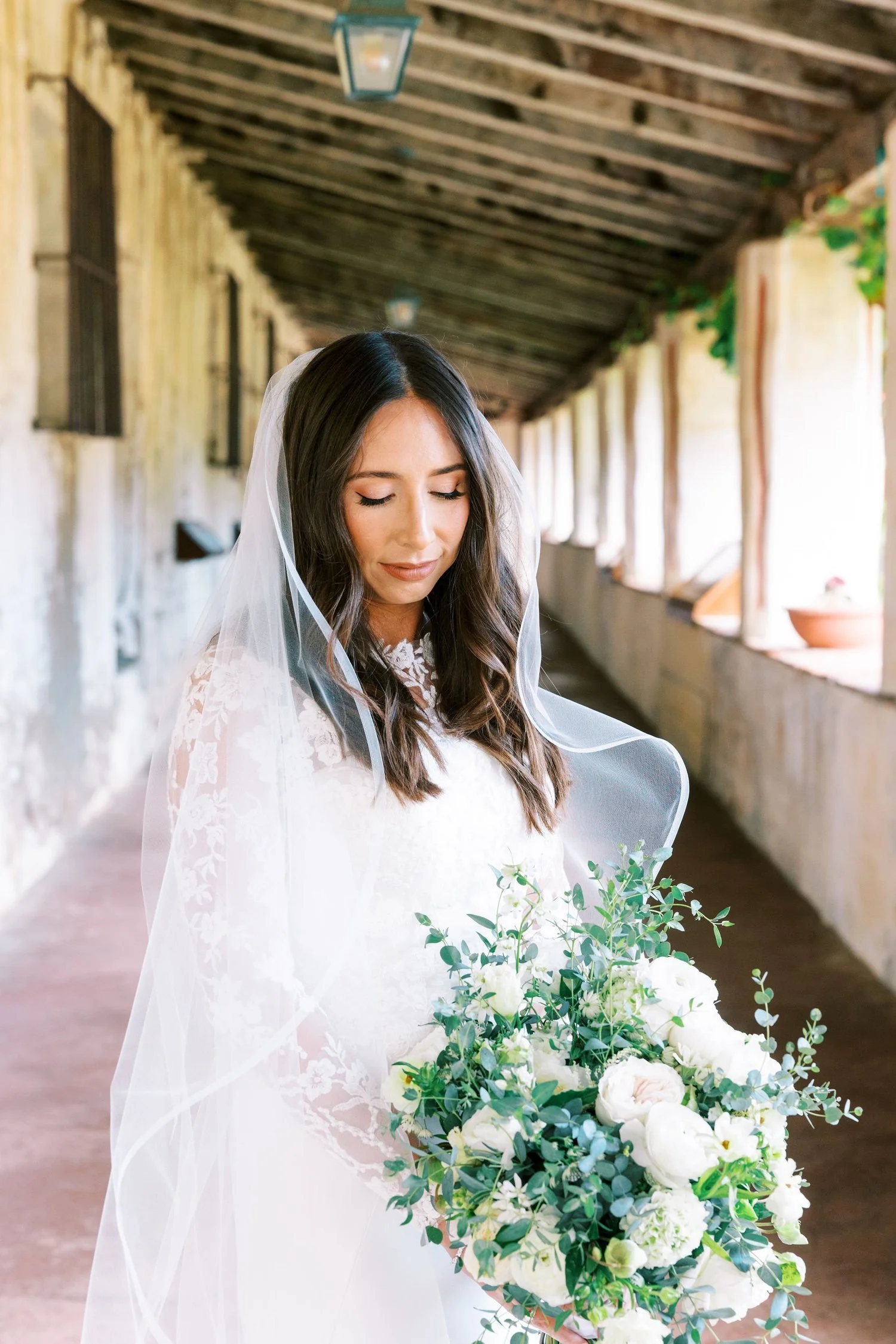 Bride in a white wedding dress with a veil, holding a bouquet of white and green flowers, standing in a rustic corridor with wooden roof and stone walls.