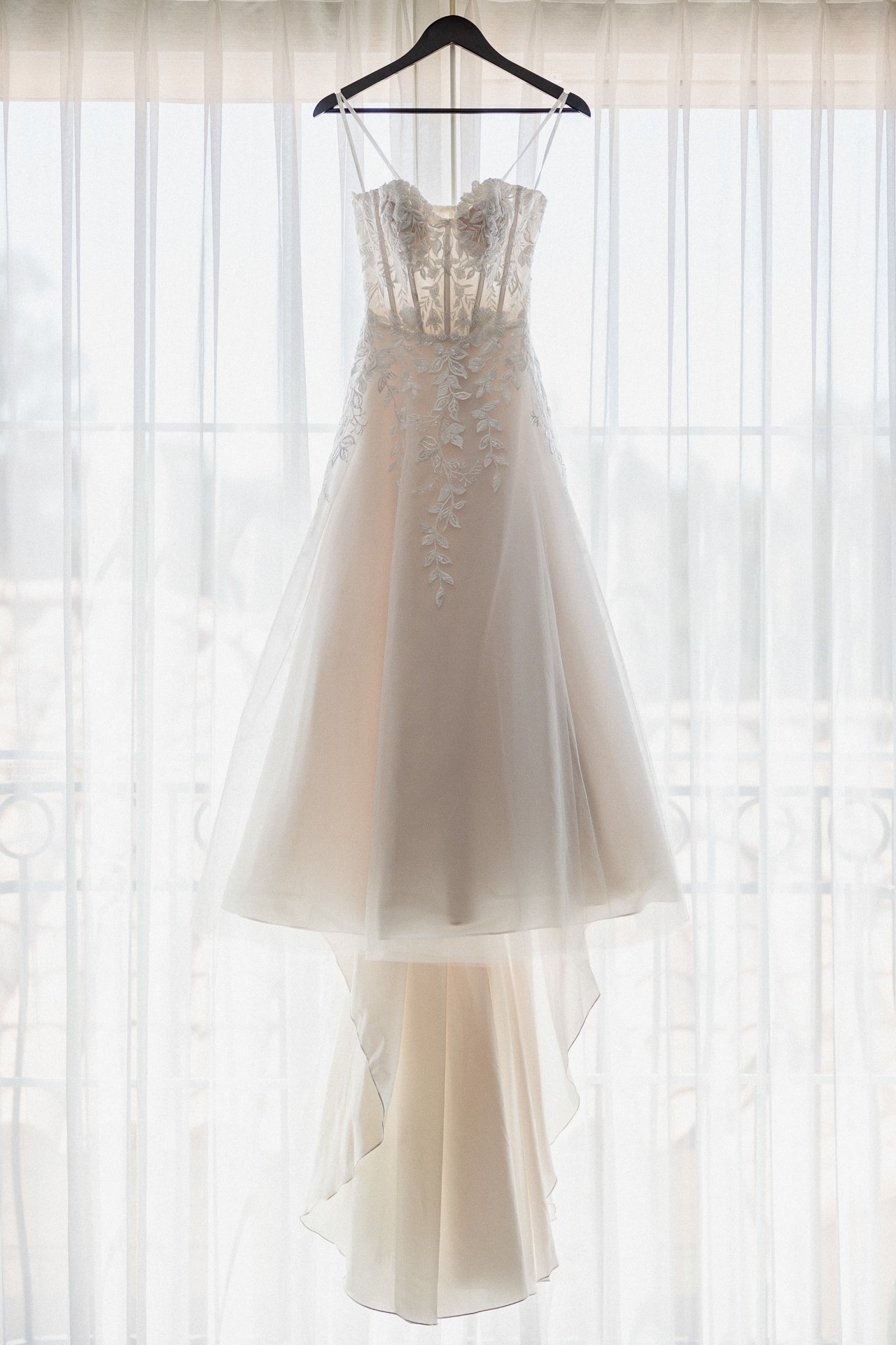 A wedding dress with lace details hanging on a black hanger in front of a window with sheer curtains.