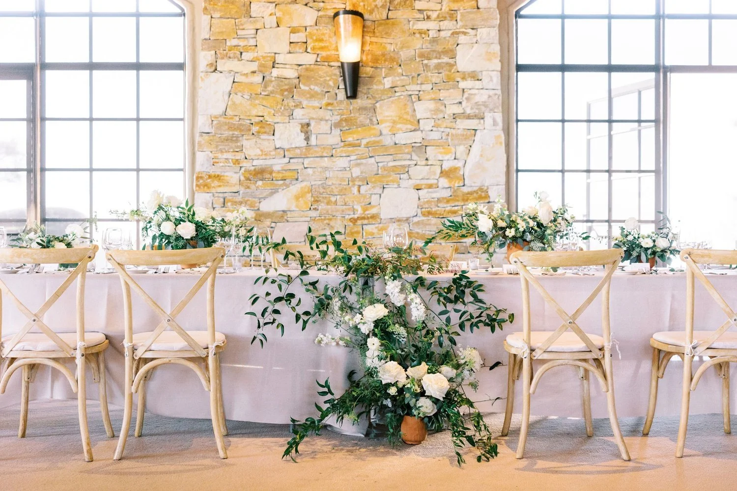 A decorated event table with floral arrangements and white chairs set against a stone wall with large windows.