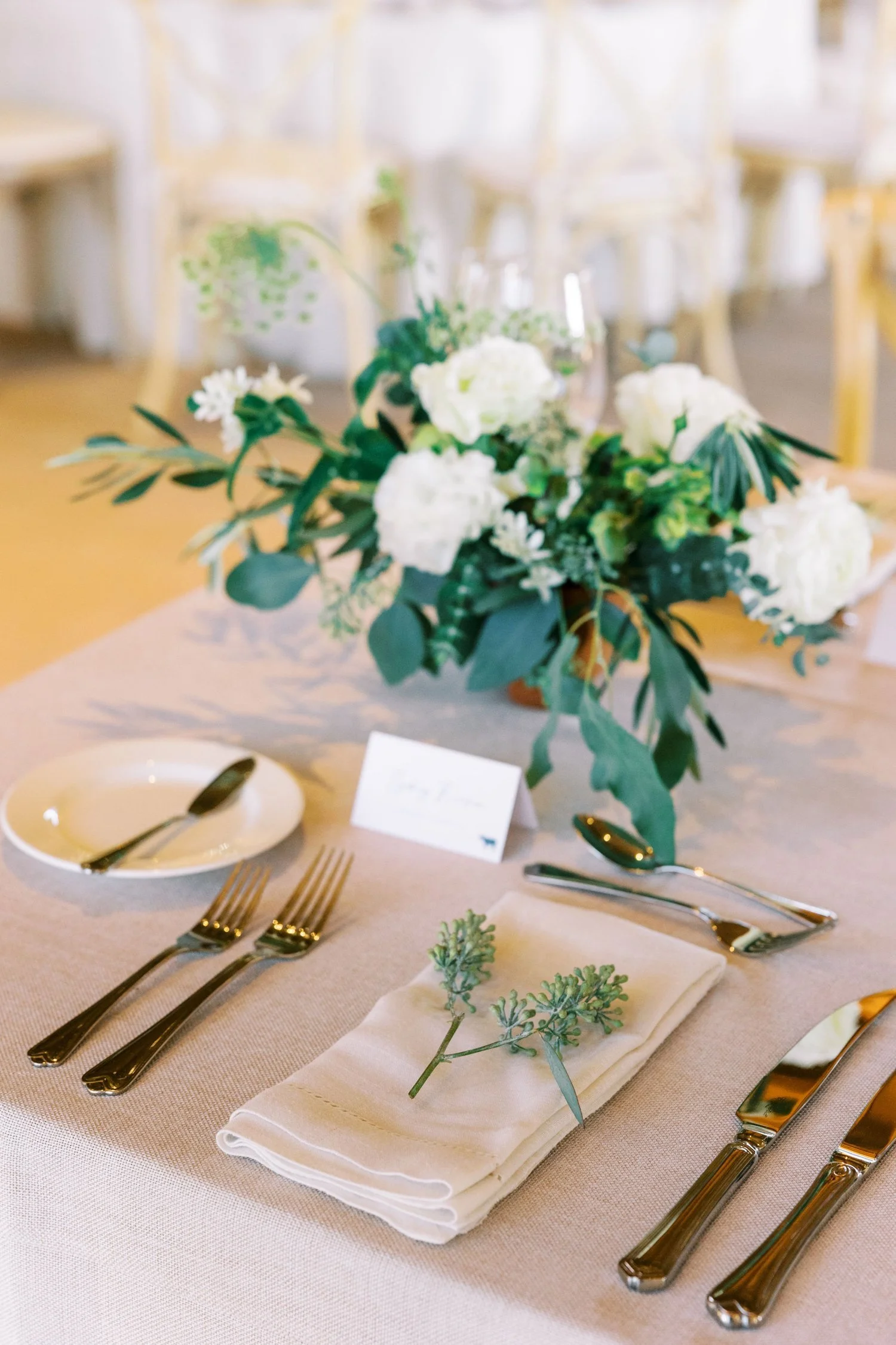Elegant table setting with a white cloth napkin folded on a beige tablecloth, topped with a sprig of greenery, silverware including forks, knives, and spoons, a small white plate with a black-handled spoon, a floral centerpiece with white flowers and