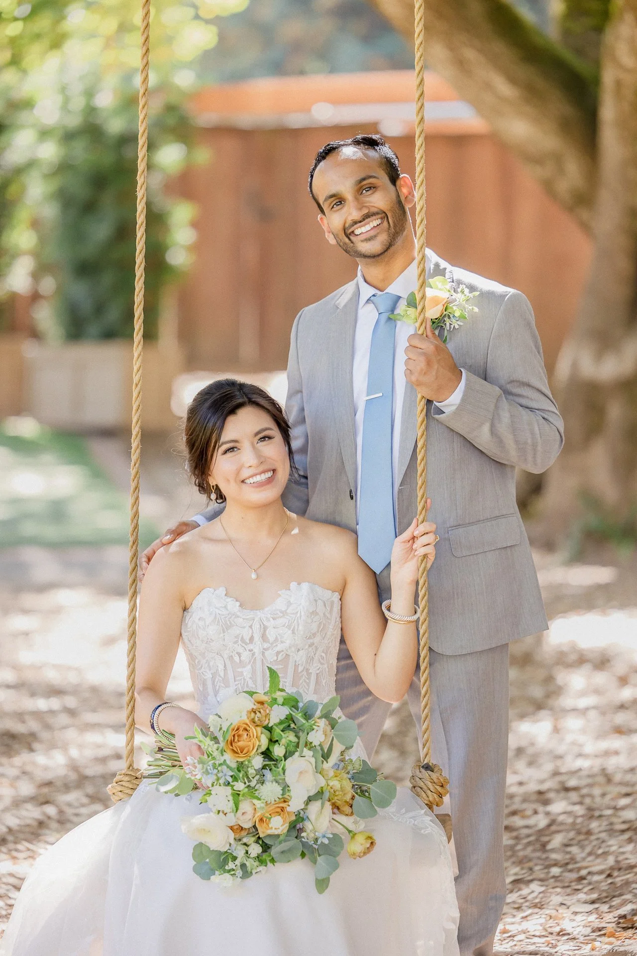 A smiling bride sitting on a swing holding a bouquet, and a groom standing beside her holding the swing's rope, outdoors surrounded by trees and sunlight.