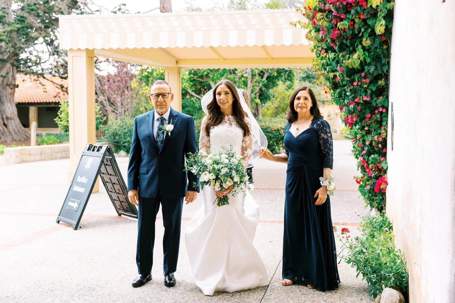 A bride in a white lace wedding dress holding a bouquet, standing between an older man in a navy suit and a woman in a dark blue dress, outdoors with greenery and pink flowers.
