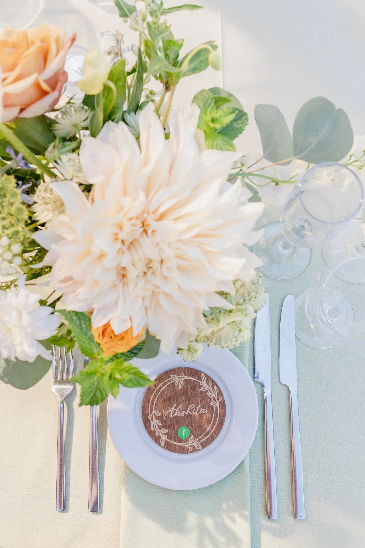 A floral centerpiece on a white plate with a name tag reading 'Aksheta,' set on a light green tablecloth. The arrangement includes large cream-colored and peach flowers, green leaves, and small white and purple blooms. Silverware, including a fork, k