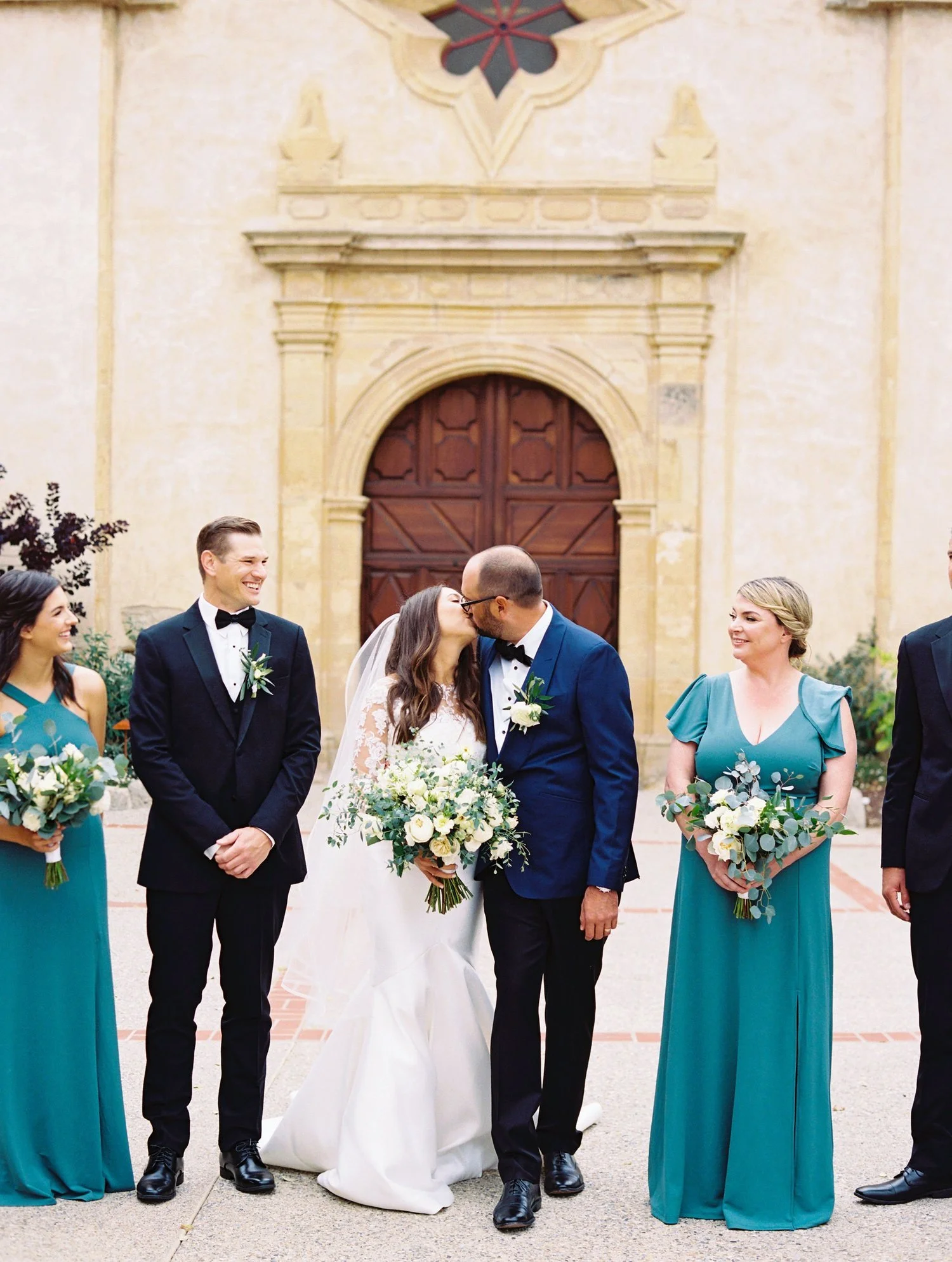 A wedding ceremony scene with the bride and groom kissing in front of an ornate arched wooden door, surrounded by bridesmaids and groomsmen holding bouquets.