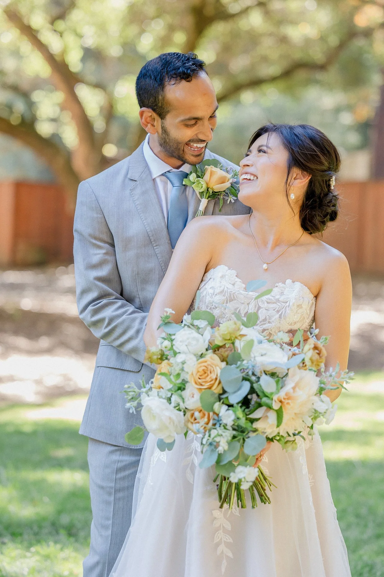 A newlywed couple smiling and sharing a joyful moment outdoors, with the bride holding a bouquet of flowers and wearing a wedding dress, while the groom in a light gray suit stands behind her.