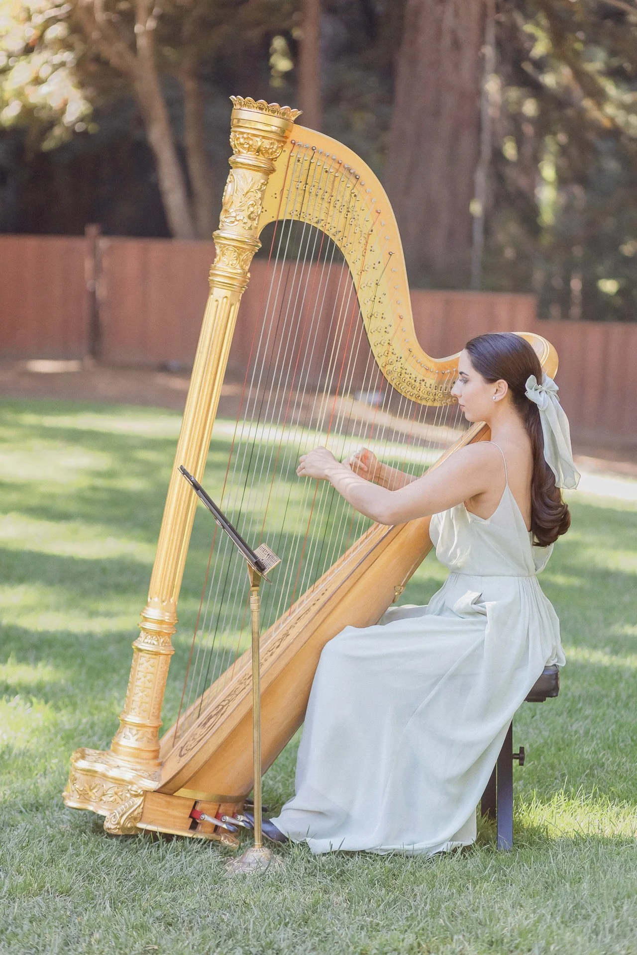 A woman in a white dress sitting on a chair outdoors, playing a large, ornate golden harp.