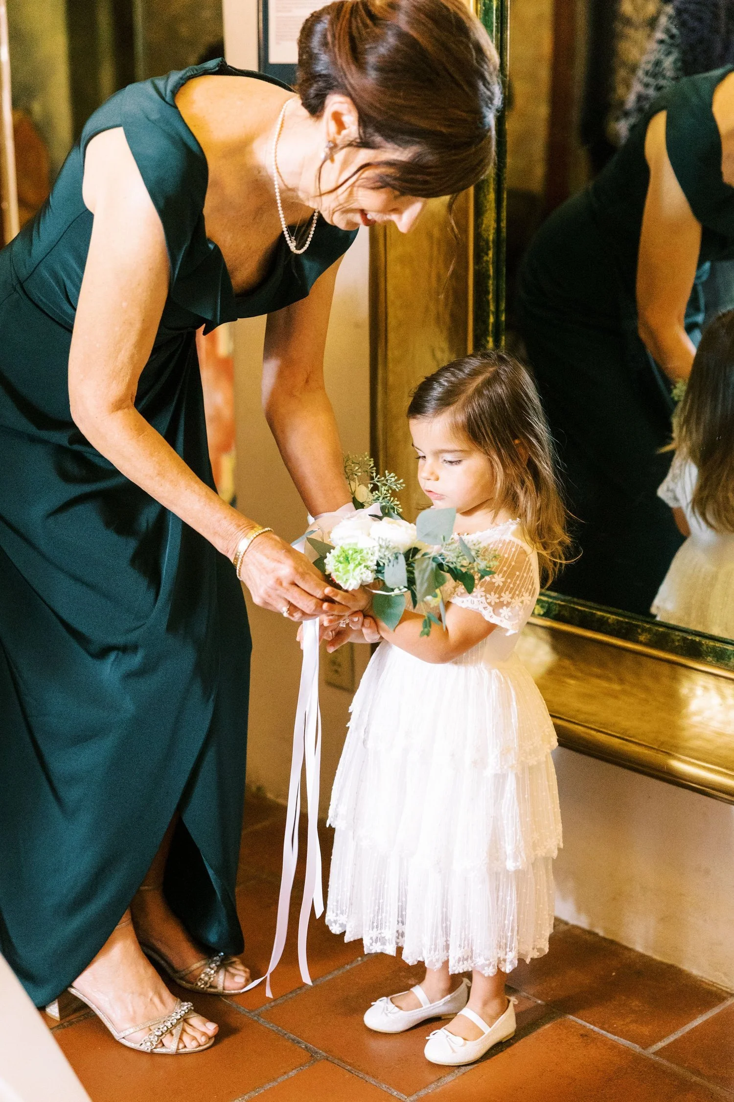 A woman in a green dress bending down to give a bouquet of flowers to a little girl in a white dress, indoors near a mirror.