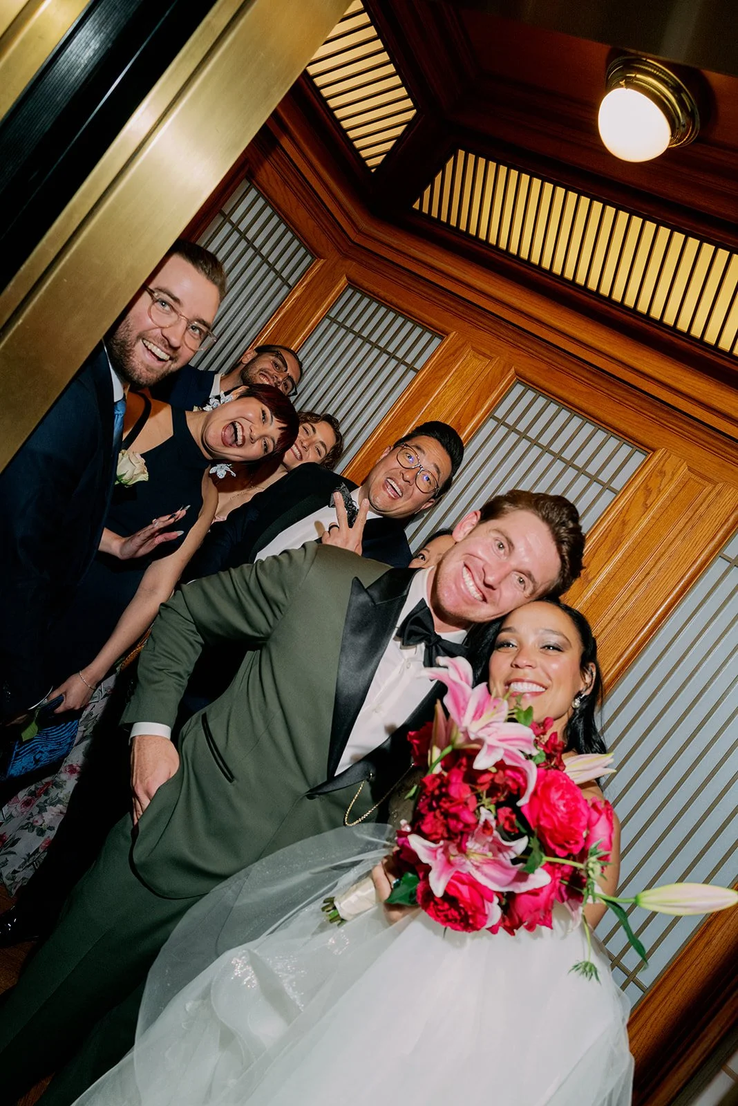 Group of wedding guests with the bride and groom in an elevator, smiling and celebrating, with the bride holding a bouquet of pink and red flowers.