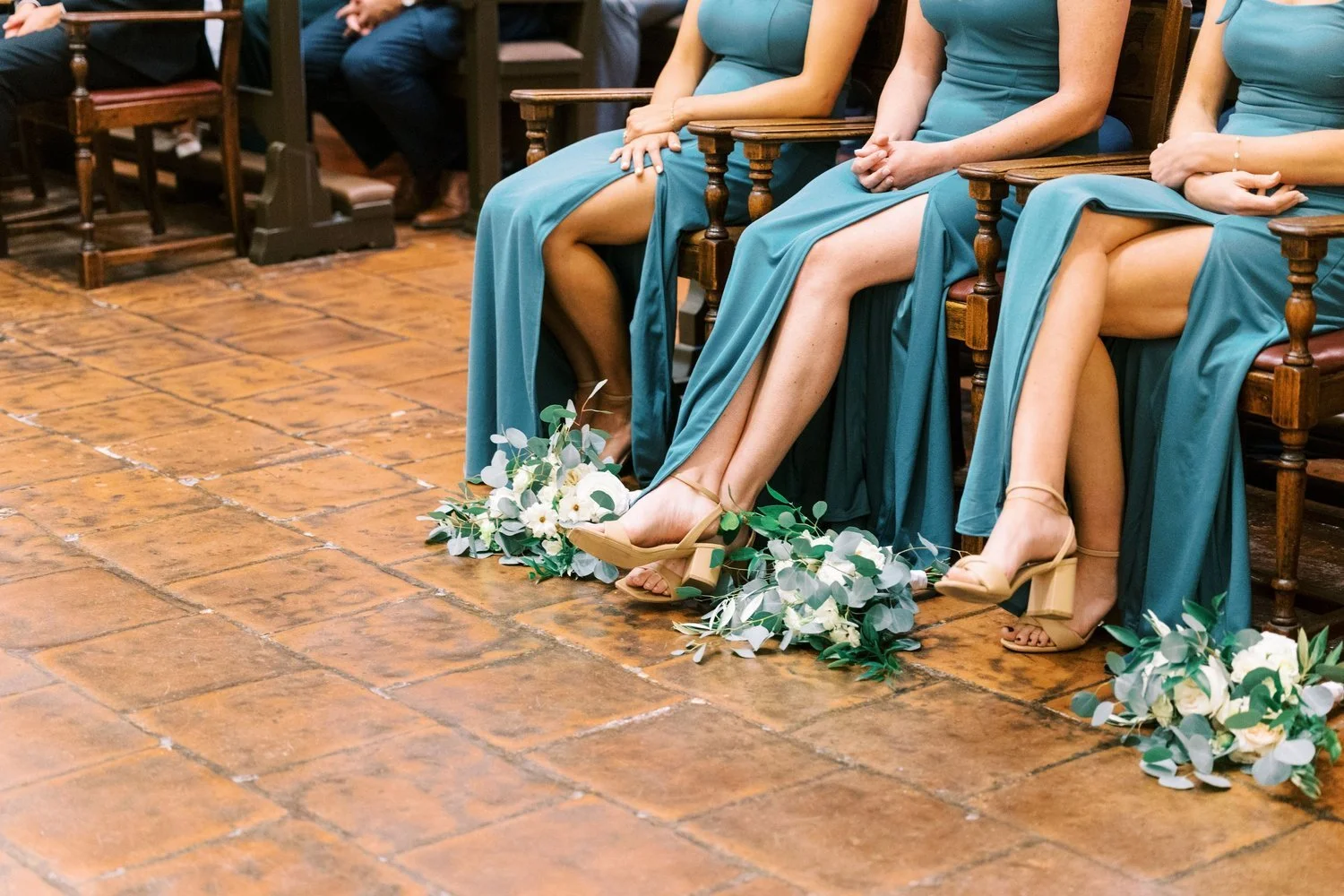 Bridesmaids sitting on chairs at a wedding ceremony, wearing teal dresses and tan heels, with bouquets of white and green flowers placed on the floor near their feet.