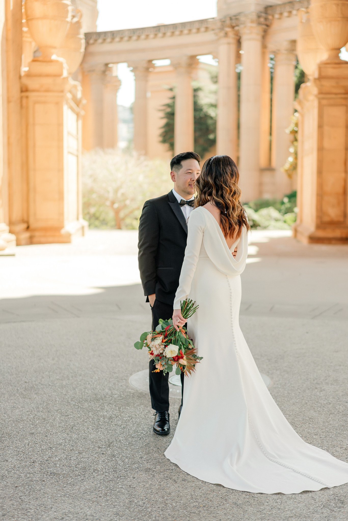 A bride and groom standing together outdoors in front of classical architecture with columns, sharing a moment and smiling at each other during their wedding.