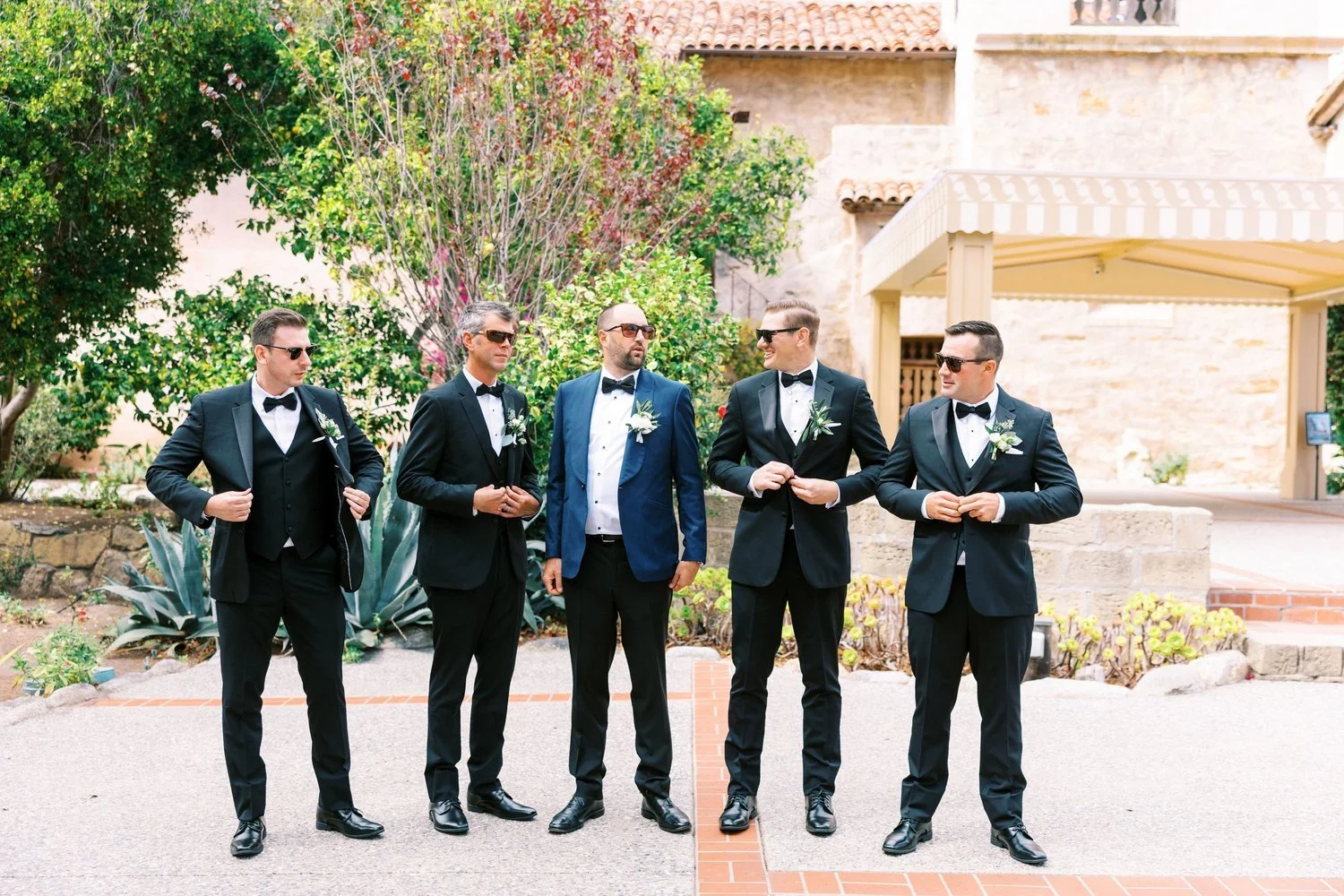 Group of five men in tuxedos with sunglasses standing outdoors in a garden during daytime.