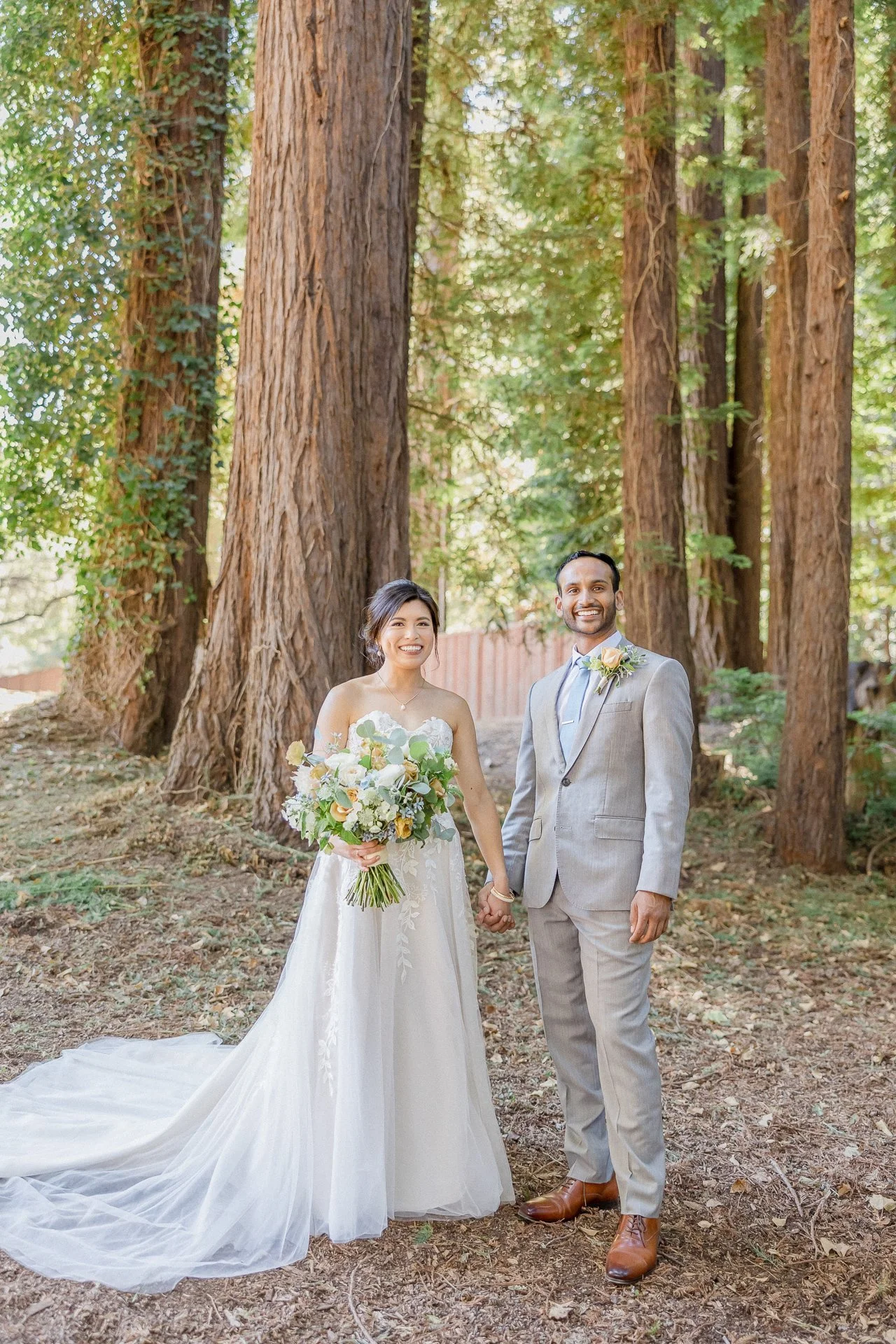 A bride and groom holding hands in a forest with tall trees, smiling at the camera, the bride wearing a white wedding dress holding a bouquet, and the groom in a light gray suit with a boutonniere.